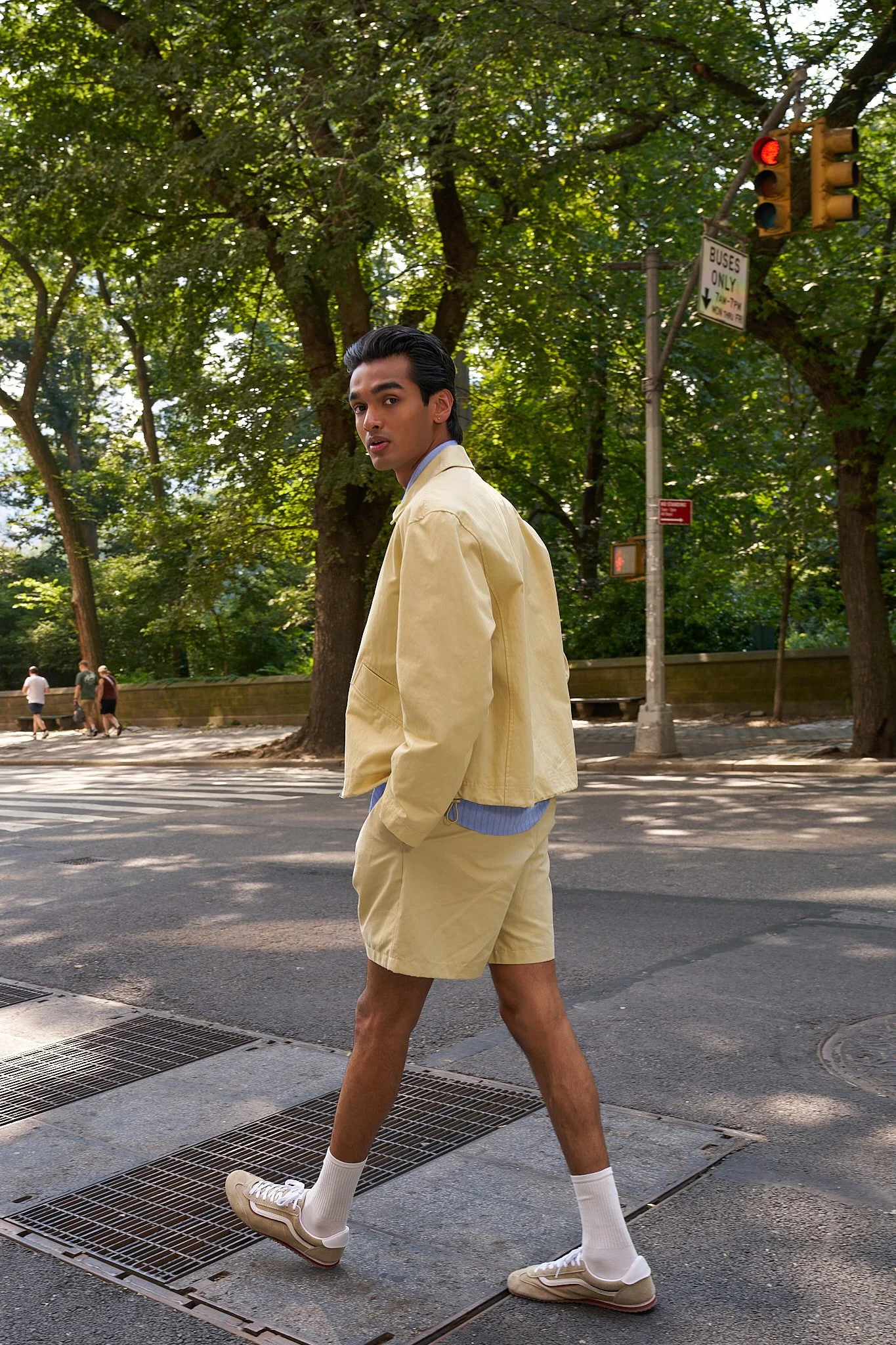 Young man with dark hair and tan skin wearing a beige jacket, matching shorts, white socks, and beige sneakers walking across a street at a pedestrian crosswalk under a tree with green leaves, with traffic lights and a 