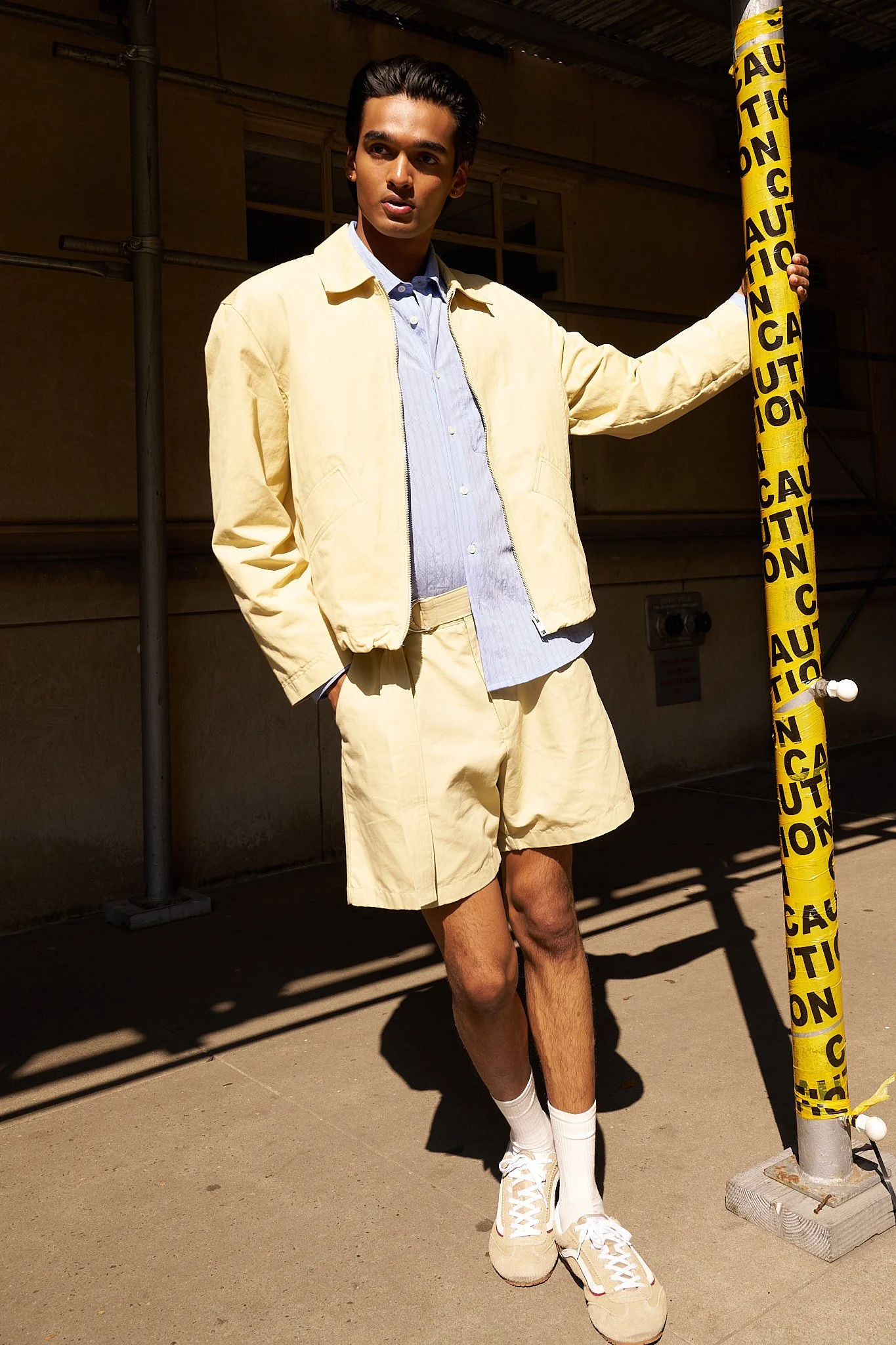 Young man standing outdoors next to a yellow caution pole, wearing a cream jacket, shorts, sneakers, and white socks, with a building in the background.