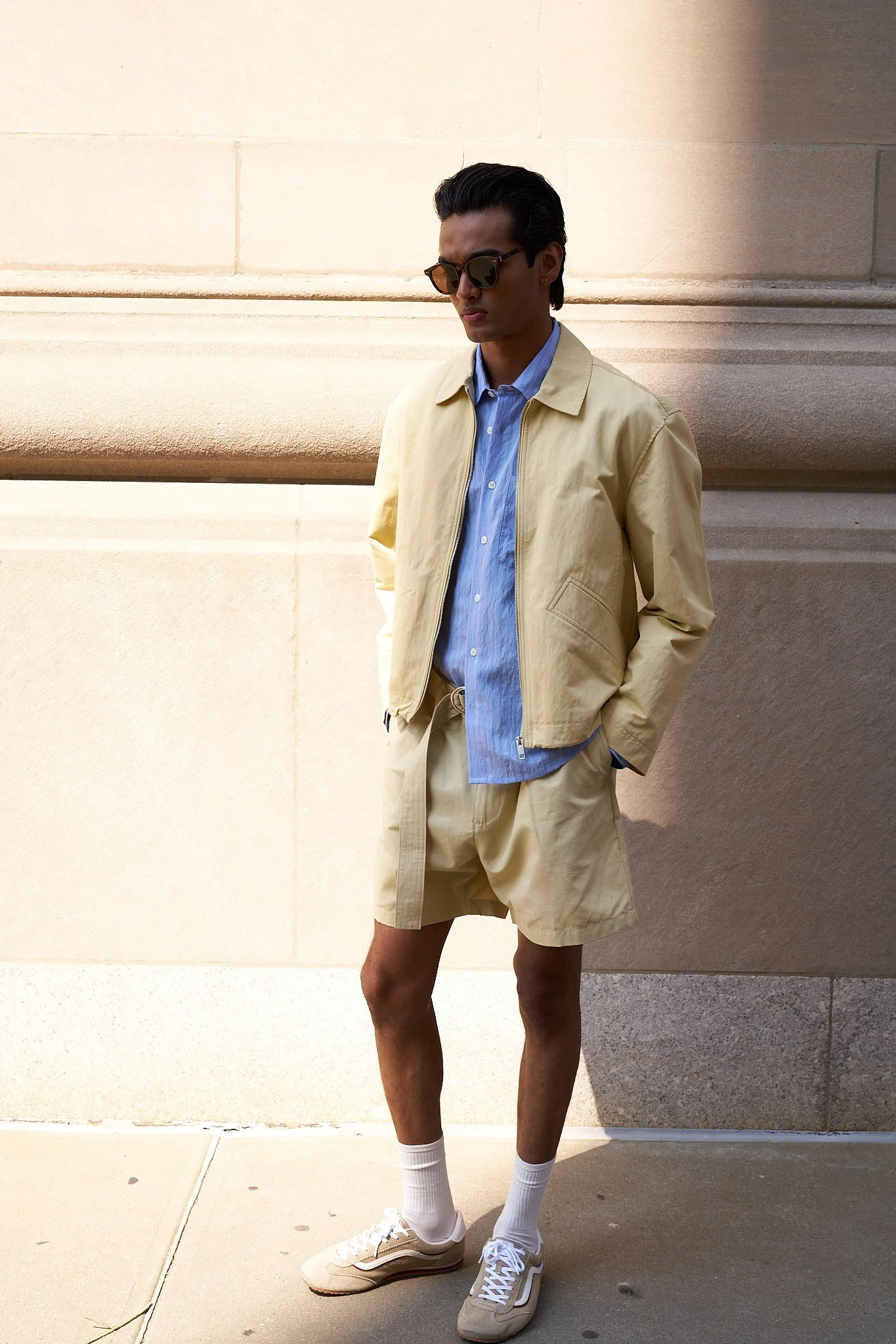 A young man in tan shorts, a blue button-up shirt, and a beige zip-up jacket stands against a beige stone wall, wearing sunglasses and beige sneakers, with his hands in his pockets and one leg slightly bent.