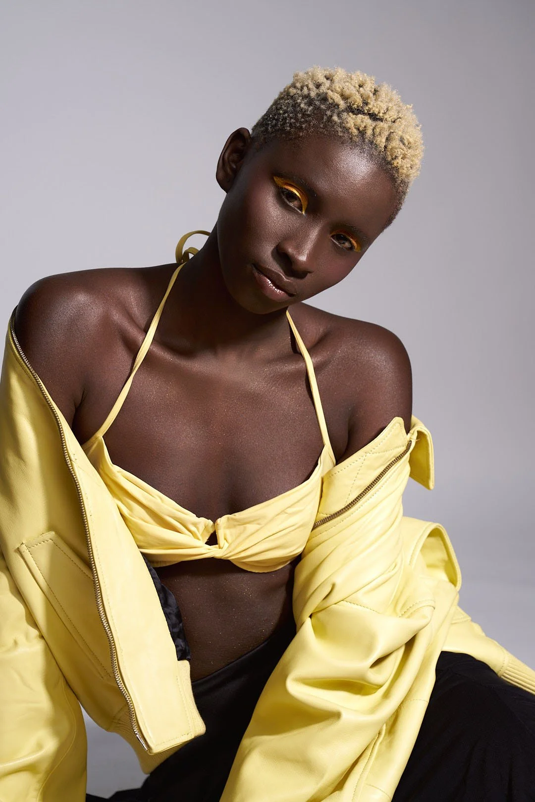 A woman with dark skin, short bleached blonde hair, yellow eyeshadow, wearing a yellow bralette, and a yellow jacket draped over her shoulders, poses against a plain grey background.