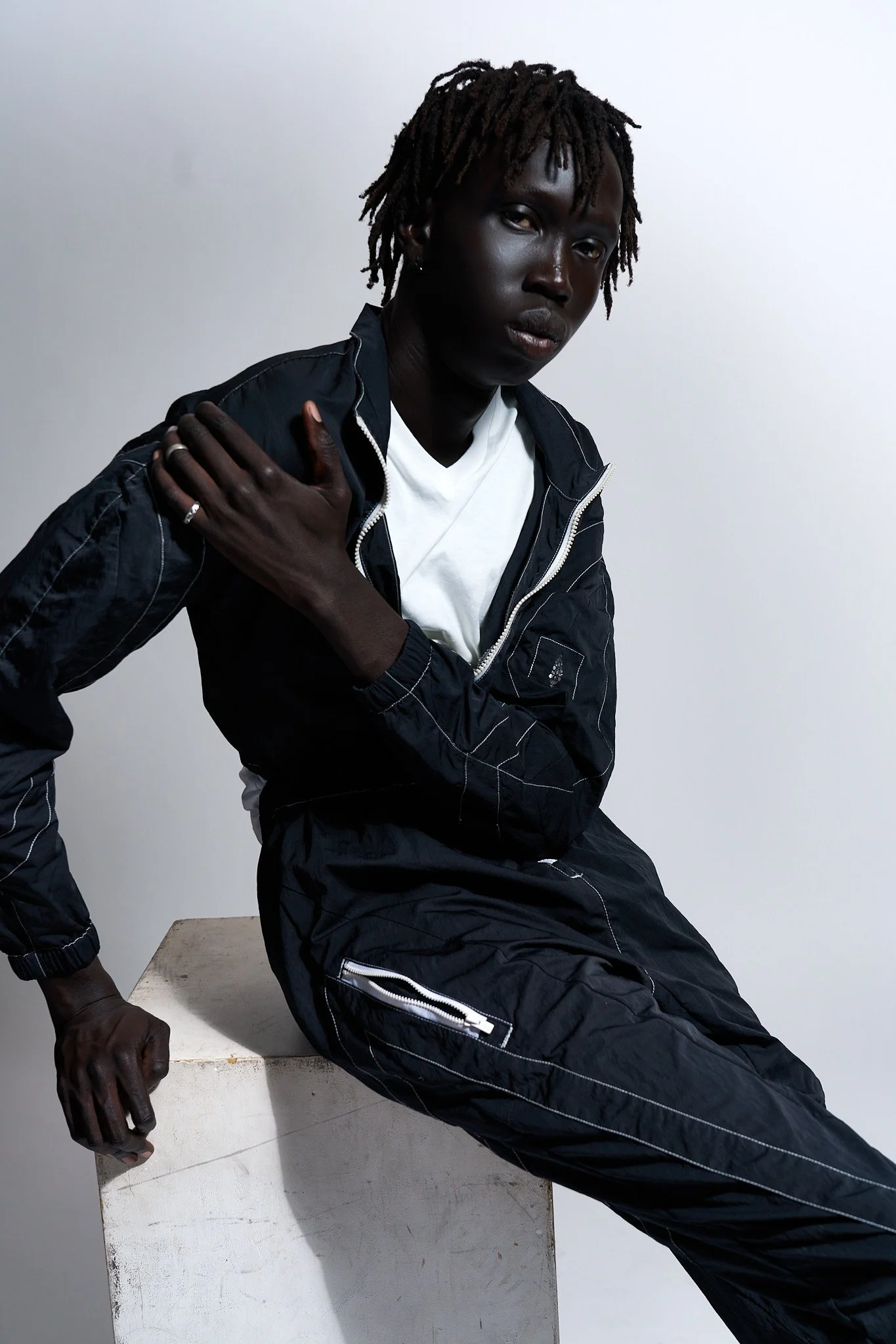 A person with dark skin and short, twisted hair, wearing a black jacket and white T-shirt, sitting on a white block against a plain light gray background, looking directly at the camera with a serious expression.
