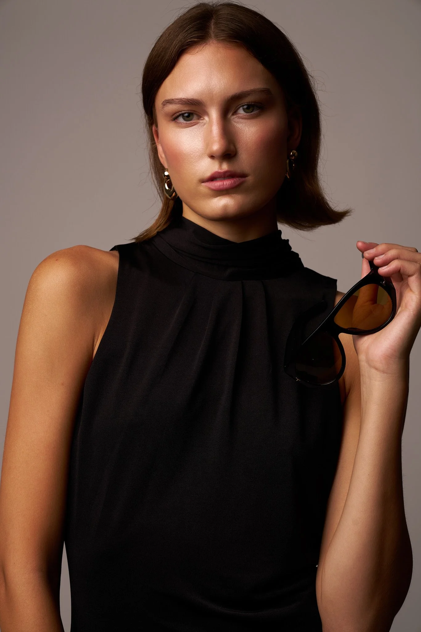 A woman with shoulder-length brown hair, wearing gold hoop earrings, a sleeveless black top with a high neck, holding a pair of large black sunglasses in her right hand, posing against a neutral gray background.