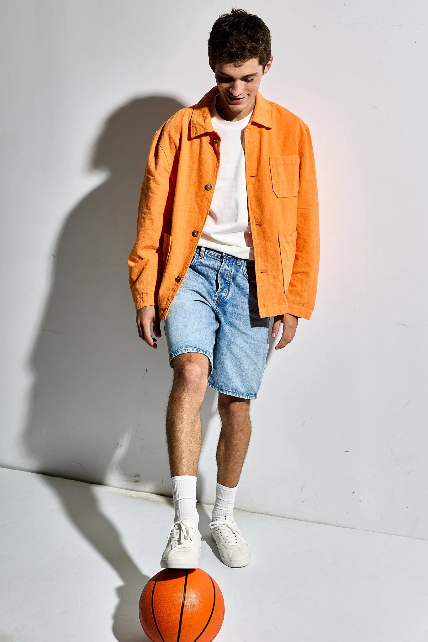 A young man with short curly hair wearing an orange jacket, white T-shirt, light blue denim shorts, white socks, and white sneakers, standing against a plain light gray background. He is balancing on a basketball with one foot, with the other foot sl