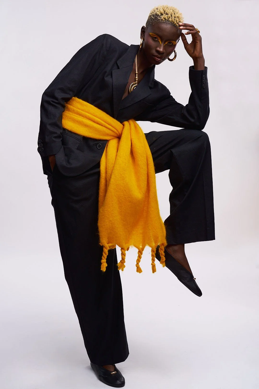 A person with short bleached blonde hair, dark skin, and bold yellow eye makeup. Wearing a black suit with a yellow scarf tied around the waist, black shoes, gold jewelry, and striking yellow eyeshadow. They are posing with one hand on their head and