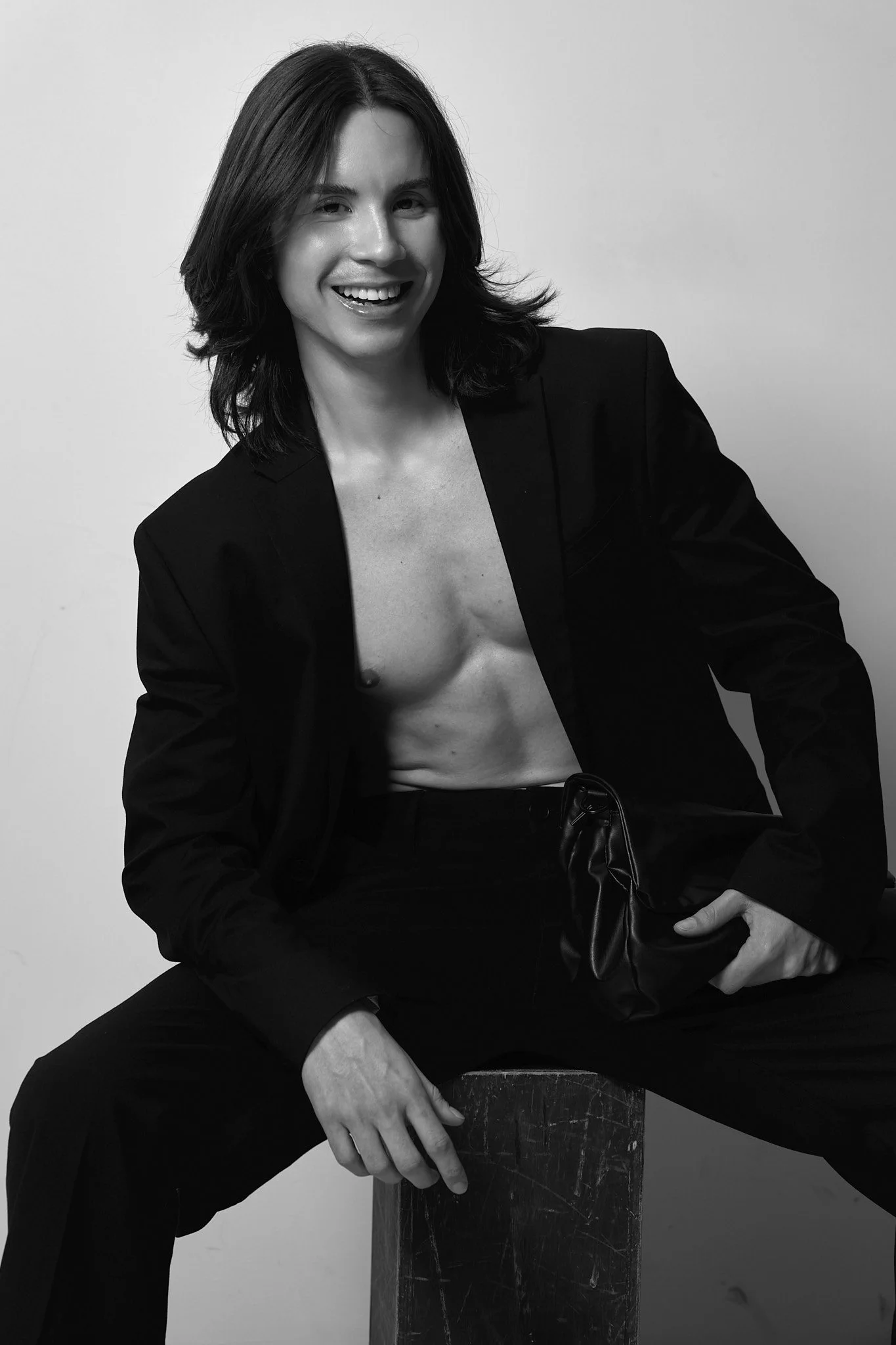 Black and white portrait of a woman with shoulder-length hair, wearing a blazer, sitting on a wooden block, and smiling.