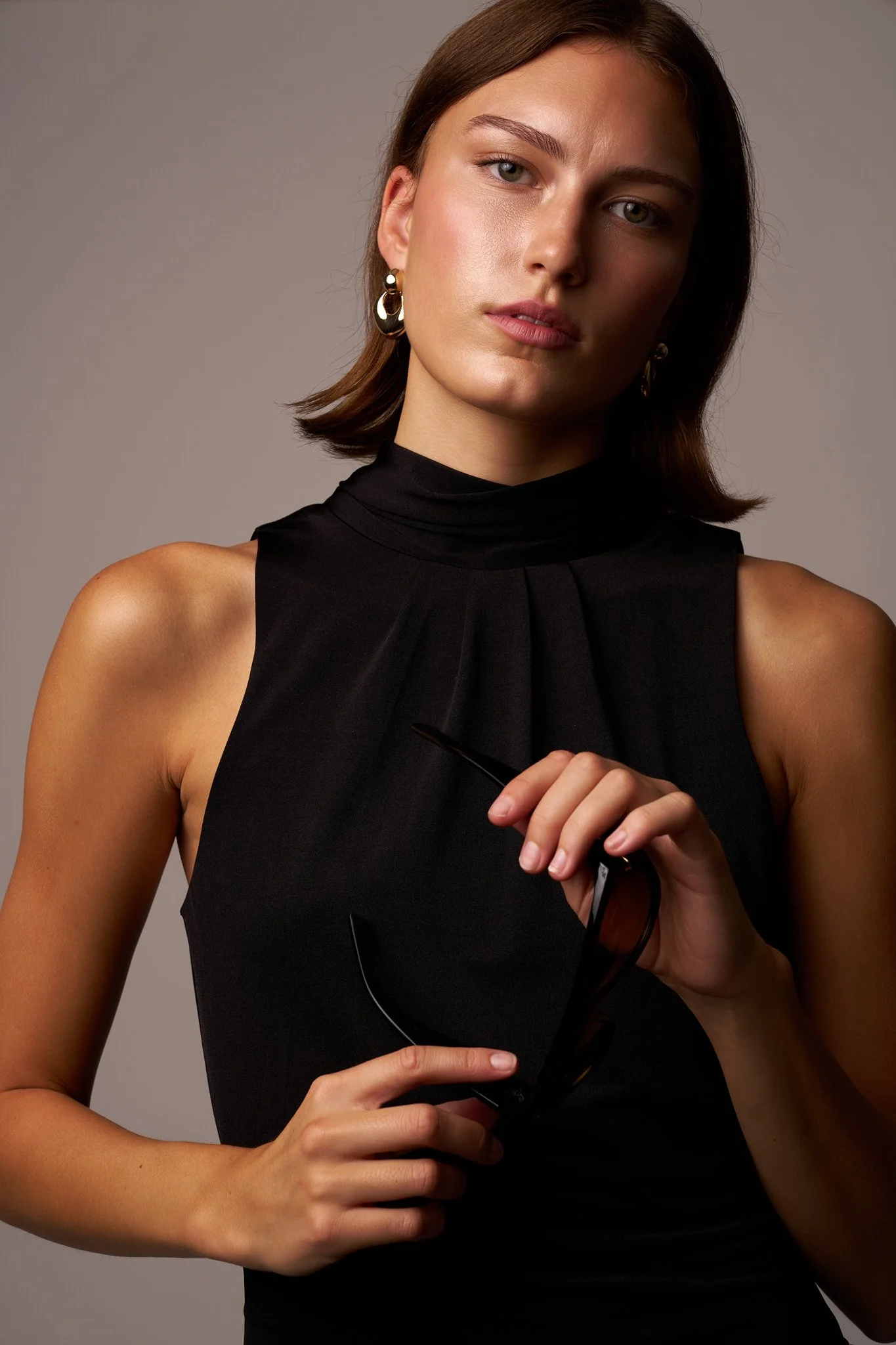A woman with shoulder-length brown hair, wearing a sleeveless black top and gold and black earrings, holding a pair of black sunglasses in her right hand against a neutral background.