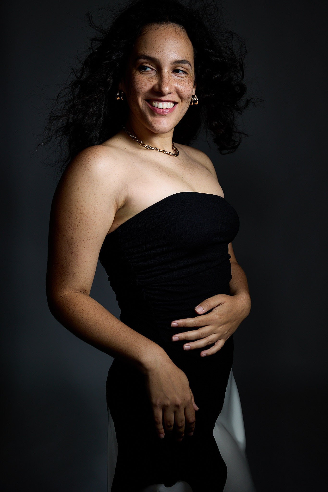 A woman with dark curly hair, light skin with freckles, and a wide smile, wearing a black strapless top and gold jewelry, posing against a dark background.