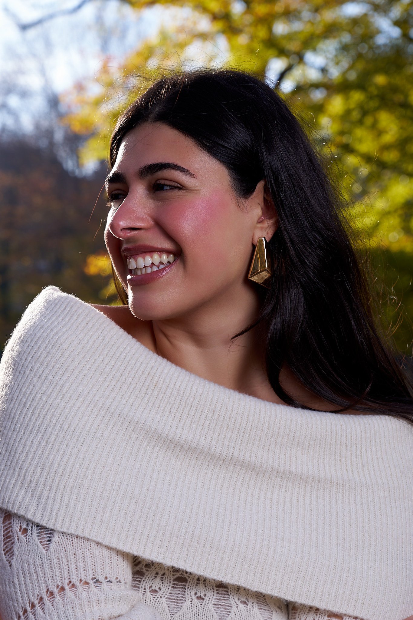 Close-up of smiling woman with dark hair, wearing a white knitted sweater and gold geometric earrings, outdoors with blurred autumn trees in the background.