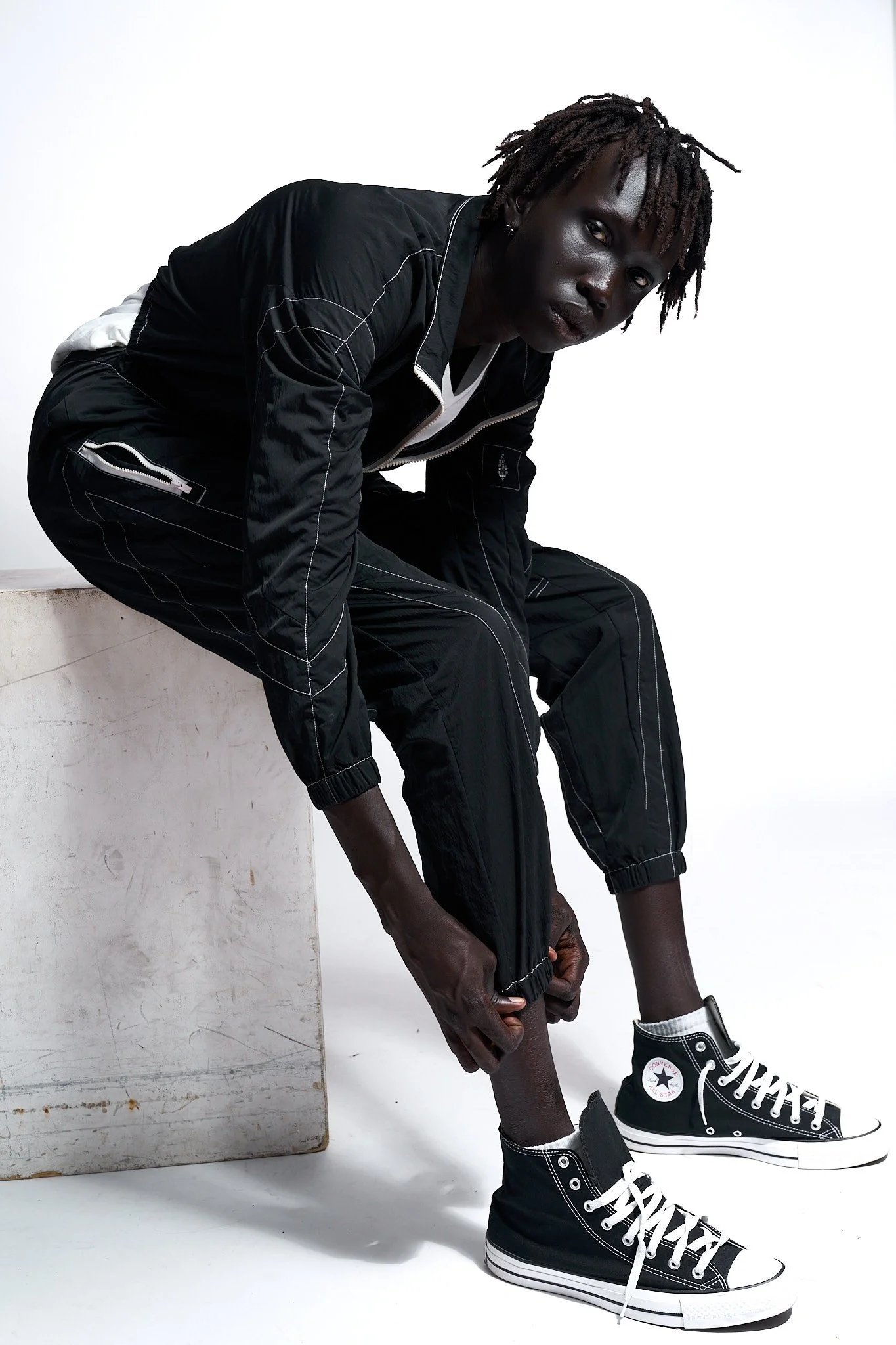 Person with dark skin and dreadlocks sitting on a white box, wearing a black jacket, black pants, and black high-top Converse sneakers, with a white background.