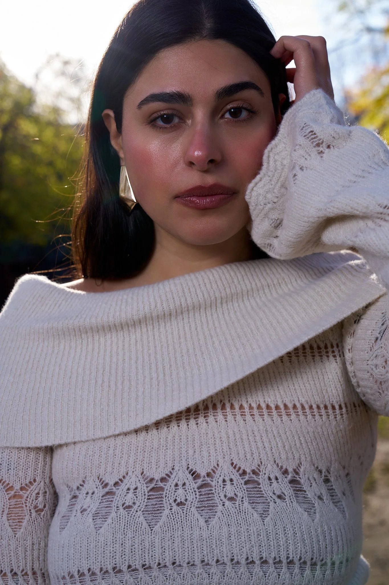 A woman with dark hair, wearing a white knitted sweater with lace-like patterns, is outdoors with blurred trees and sky in the background. She is looking directly at the camera with her right hand touching her hair.