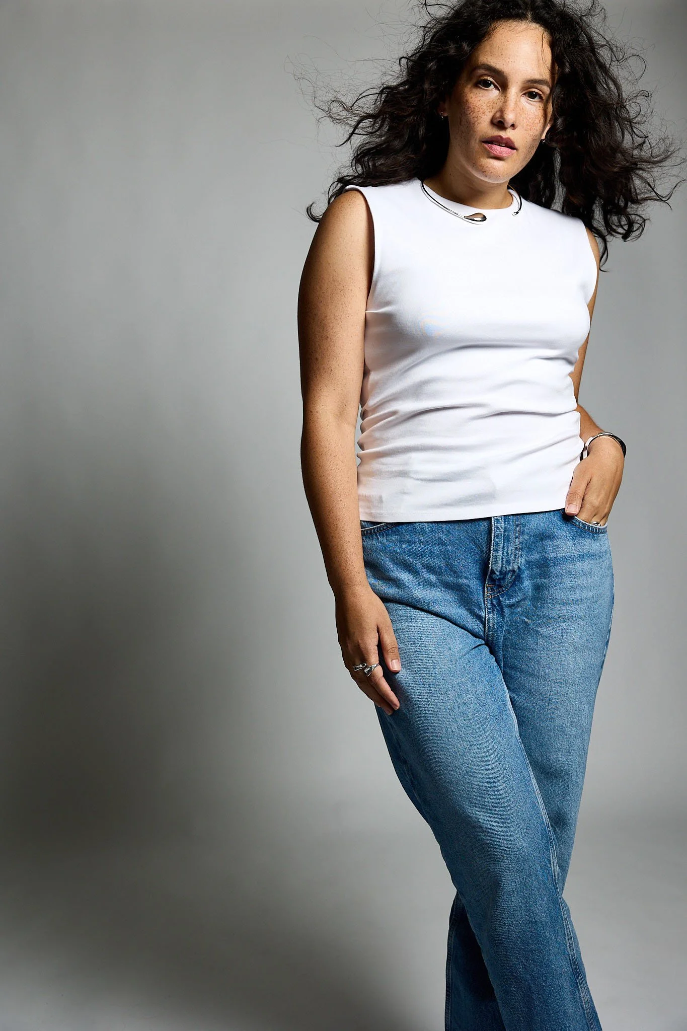 A young woman with curly dark hair, freckles, and light skin, wearing a white sleeveless top, blue jeans, and a smartwatch, standing against a gray background.