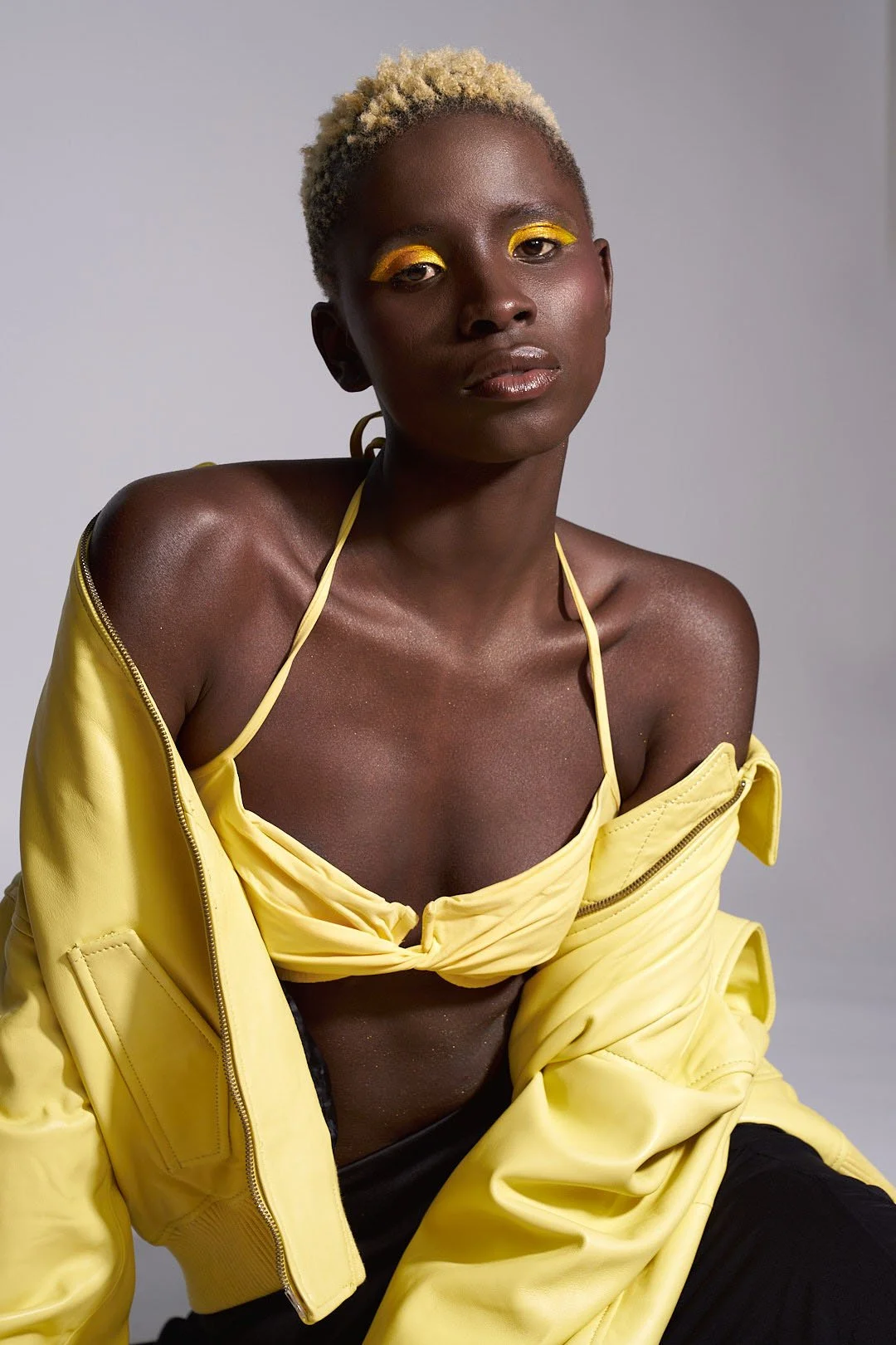 A woman with short, bleached blonde hair and dark skin wearing a yellow jacket partially off her shoulders, a yellow bra, and yellow eyeshadow.