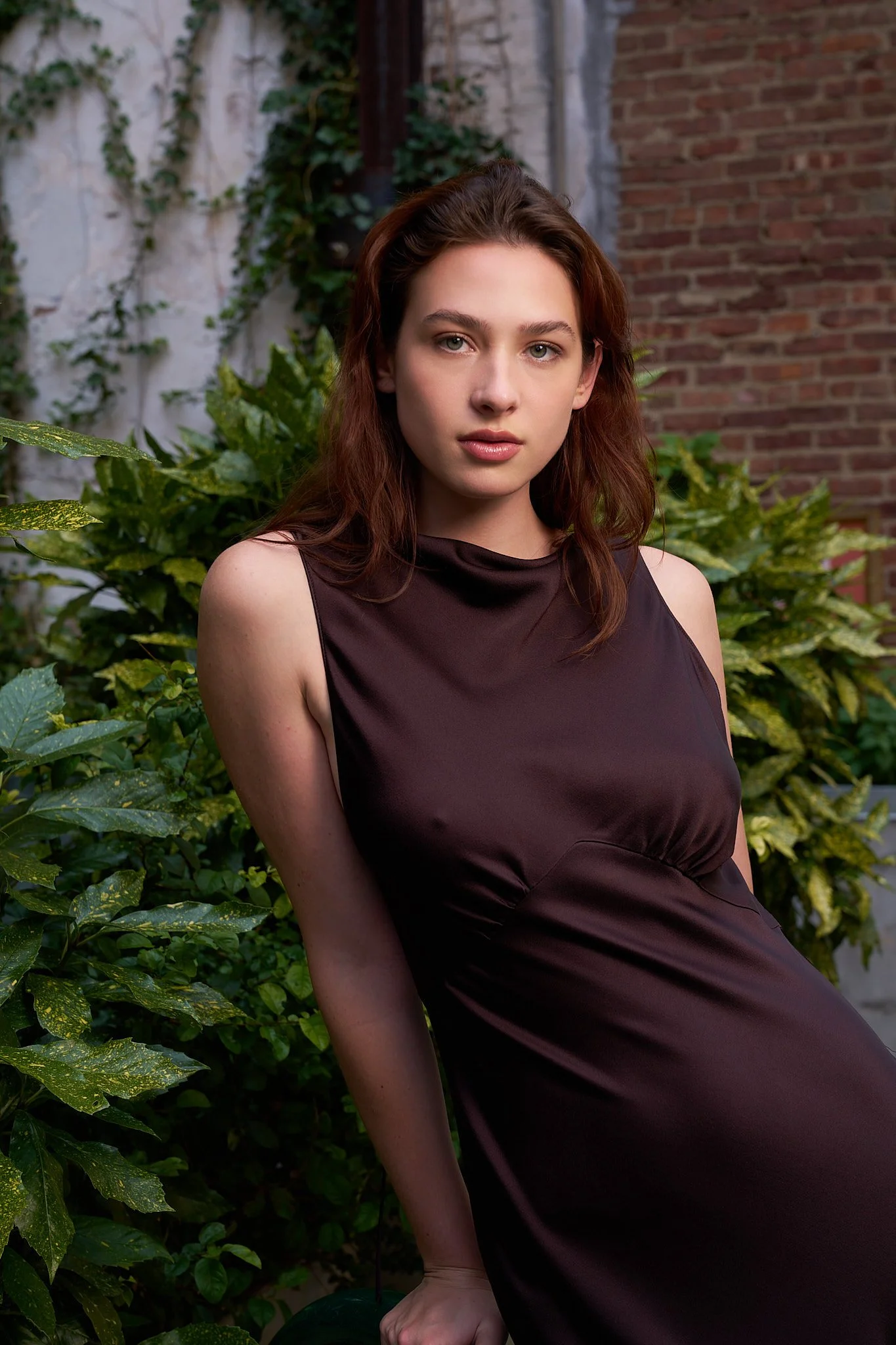 A woman with auburn hair and light skin poses in front of green foliage and a brick wall, wearing a sleeveless dark purple dress.
