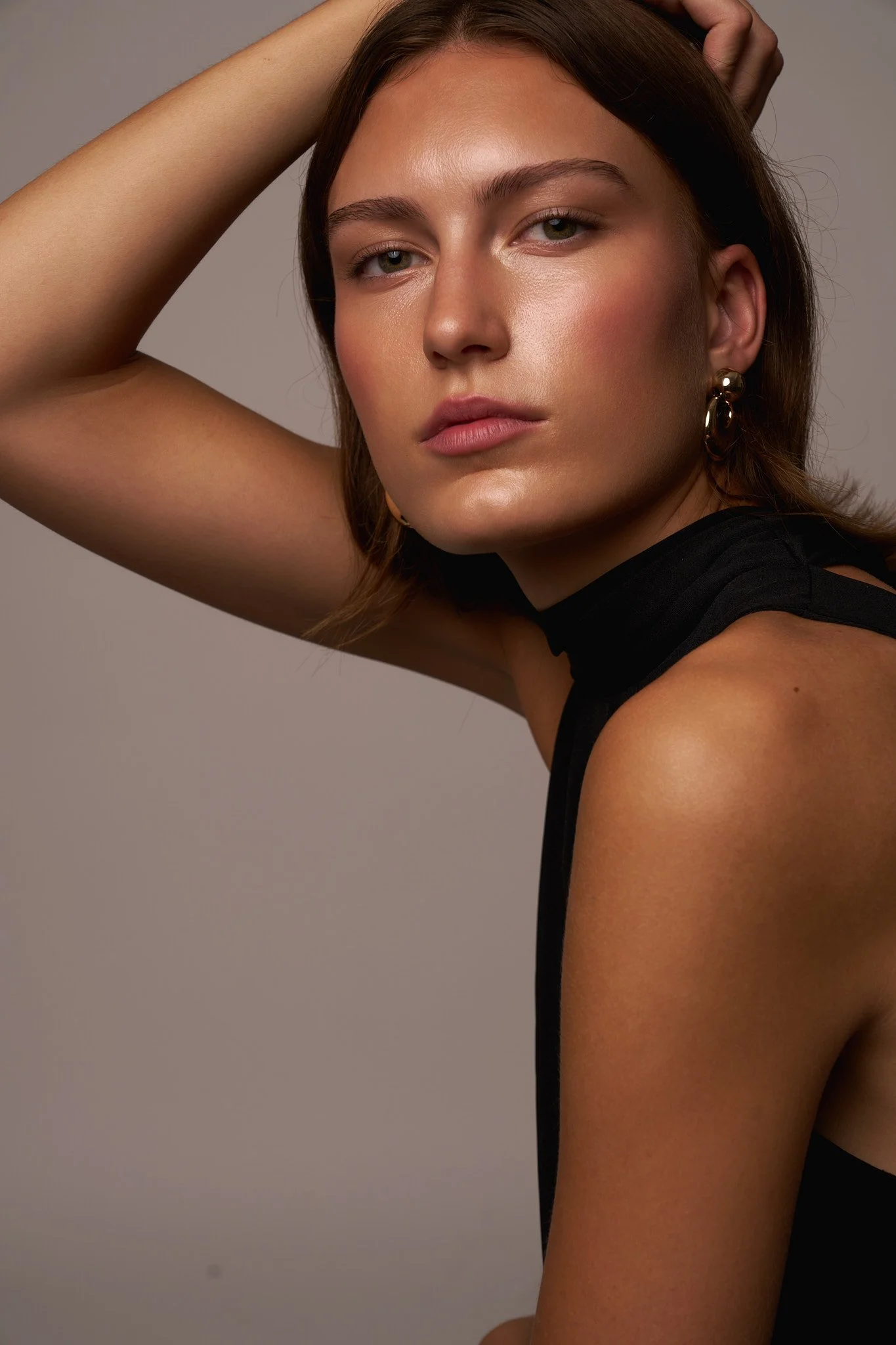 Close-up portrait of a woman with fair skin and brown hair, wearing a black sleeveless top and gold earrings, posing with her right arm raised and hand resting on her head, against a neutral background.