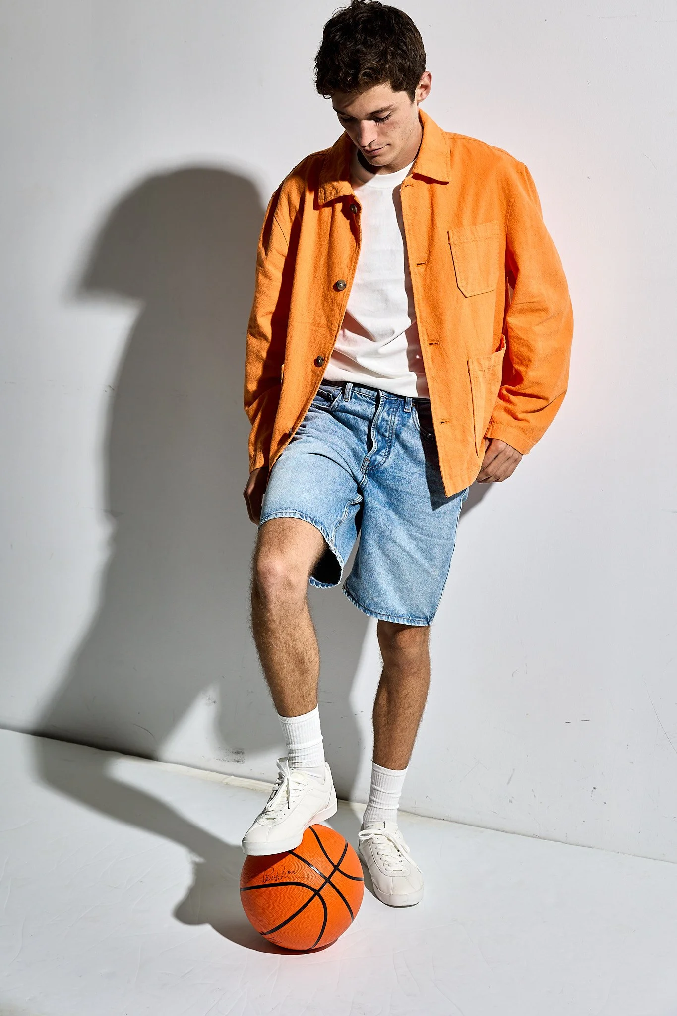 A young man with dark curly hair wearing an orange jacket, white T-shirt, denim shorts, white socks, and white sneakers standing against a plain white background. His left foot is placed on a basketball, and he is looking down at it. The shadow of hi