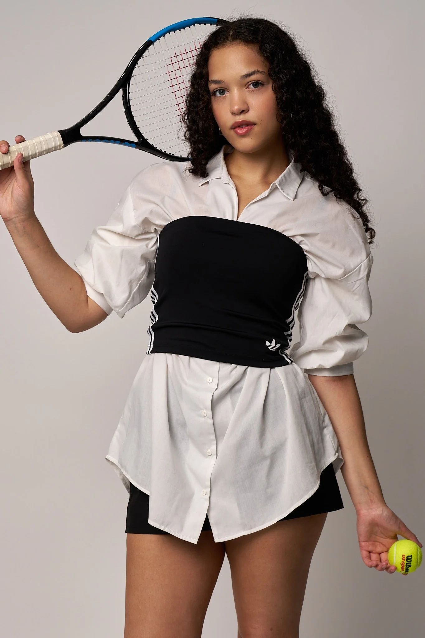 Woman with curly hair holding a tennis racket over her shoulder and a tennis ball in hand, wearing a white shirt, black Adidas shorts, and a black sports top.