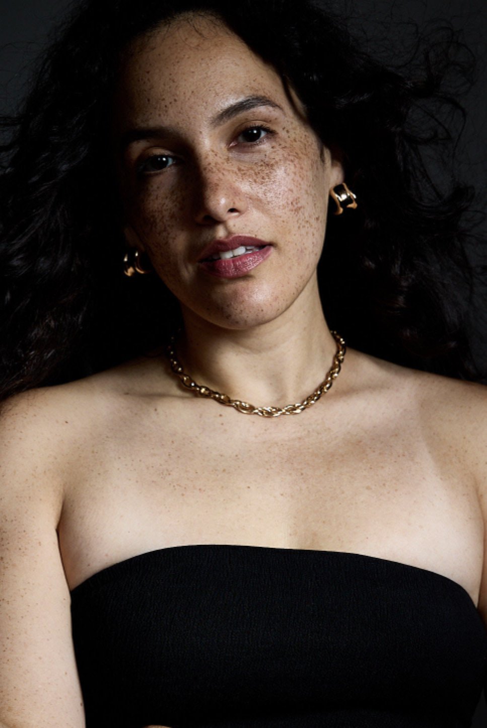 A woman with curly dark hair, wearing a black strapless top, gold jewelry including earrings, a chain necklace, and a bracelet. She has light skin with freckles and is looking directly at the camera against a black background.