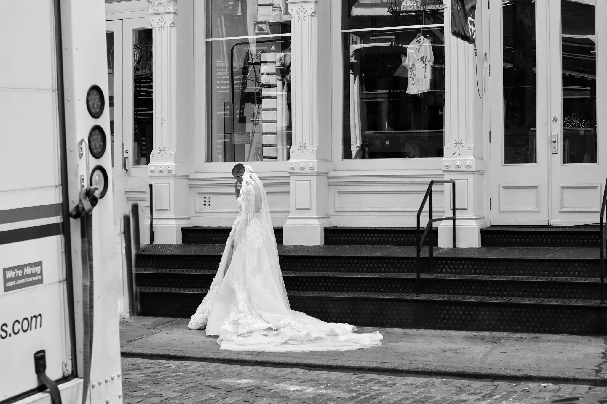 A black-and-white photo of a bride in a wedding dress standing on steps outside a building with large front windows and decorative architectural details. She appears to be adjusting her veil or grooming herself.