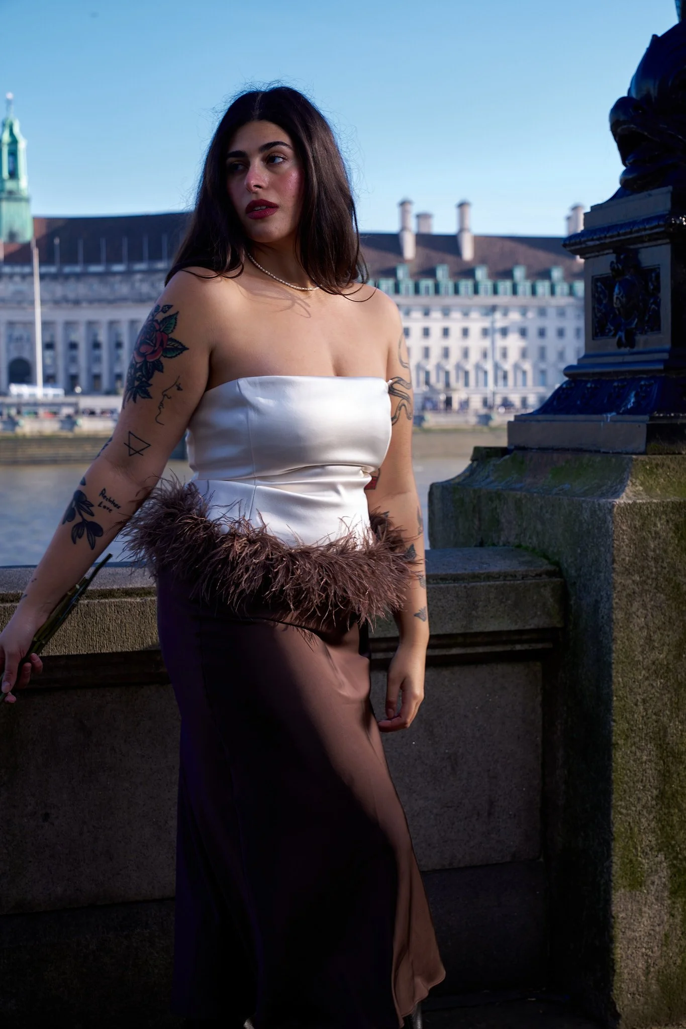 A woman with dark hair and tattoos on her arms, wearing a white strapless top with a brown skirt, holding a fan, standing near a river with classical buildings and a church with a green dome in the background.