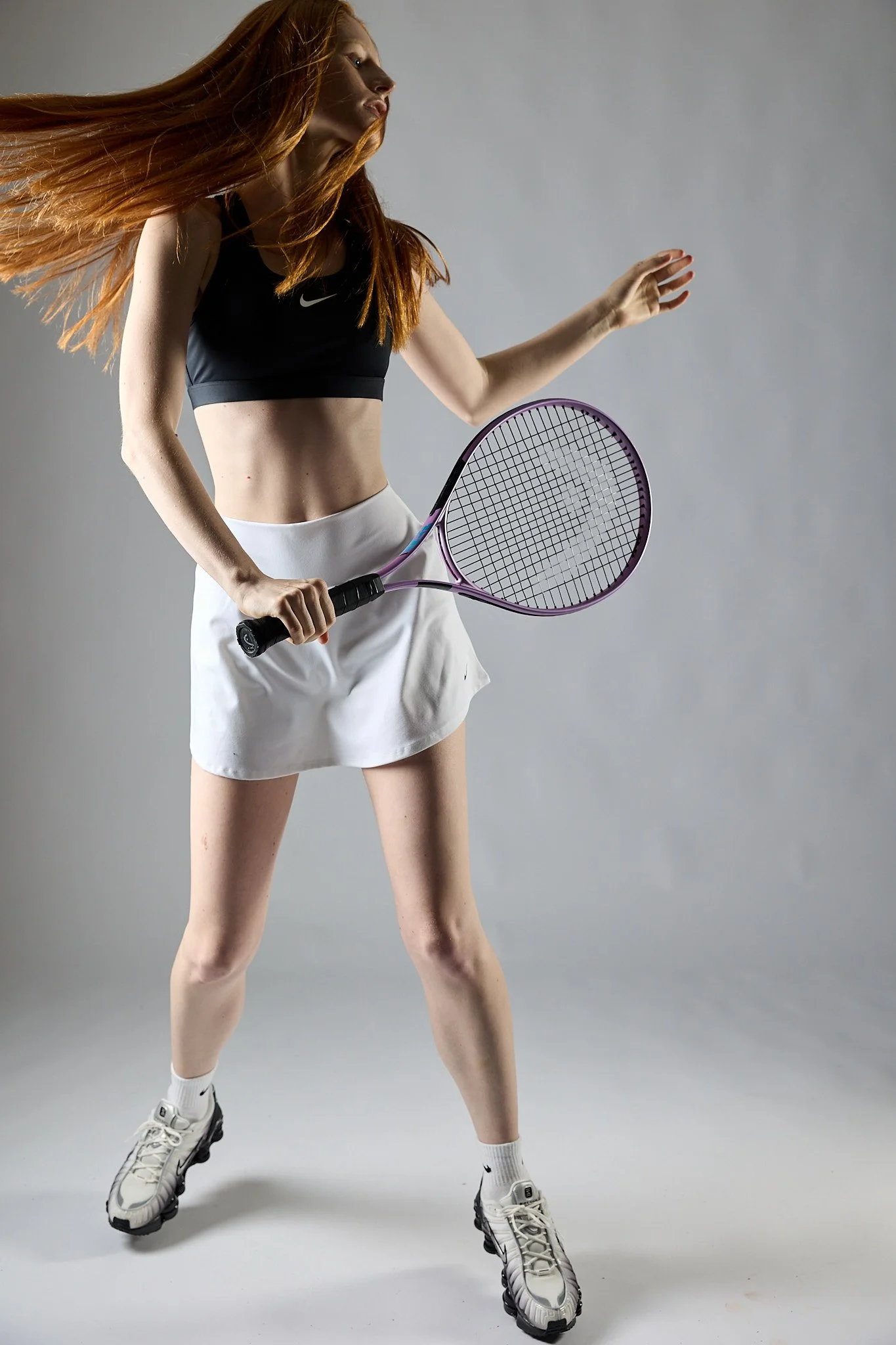 A young woman with long red hair wearing a black sports bra, white skirt, white Nike socks, and white athletic shoes, holding a tennis racket. She is in a ready stance against a plain gray background.