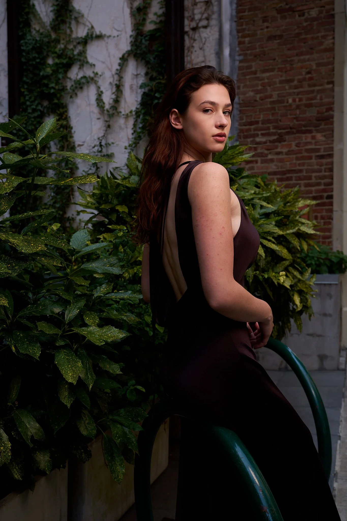 A young woman with long, wavy brown hair and fair skin has a serious expression while looking over her shoulder. She is wearing a sleeveless dress and standing outdoors near lush green plants and a red brick wall.