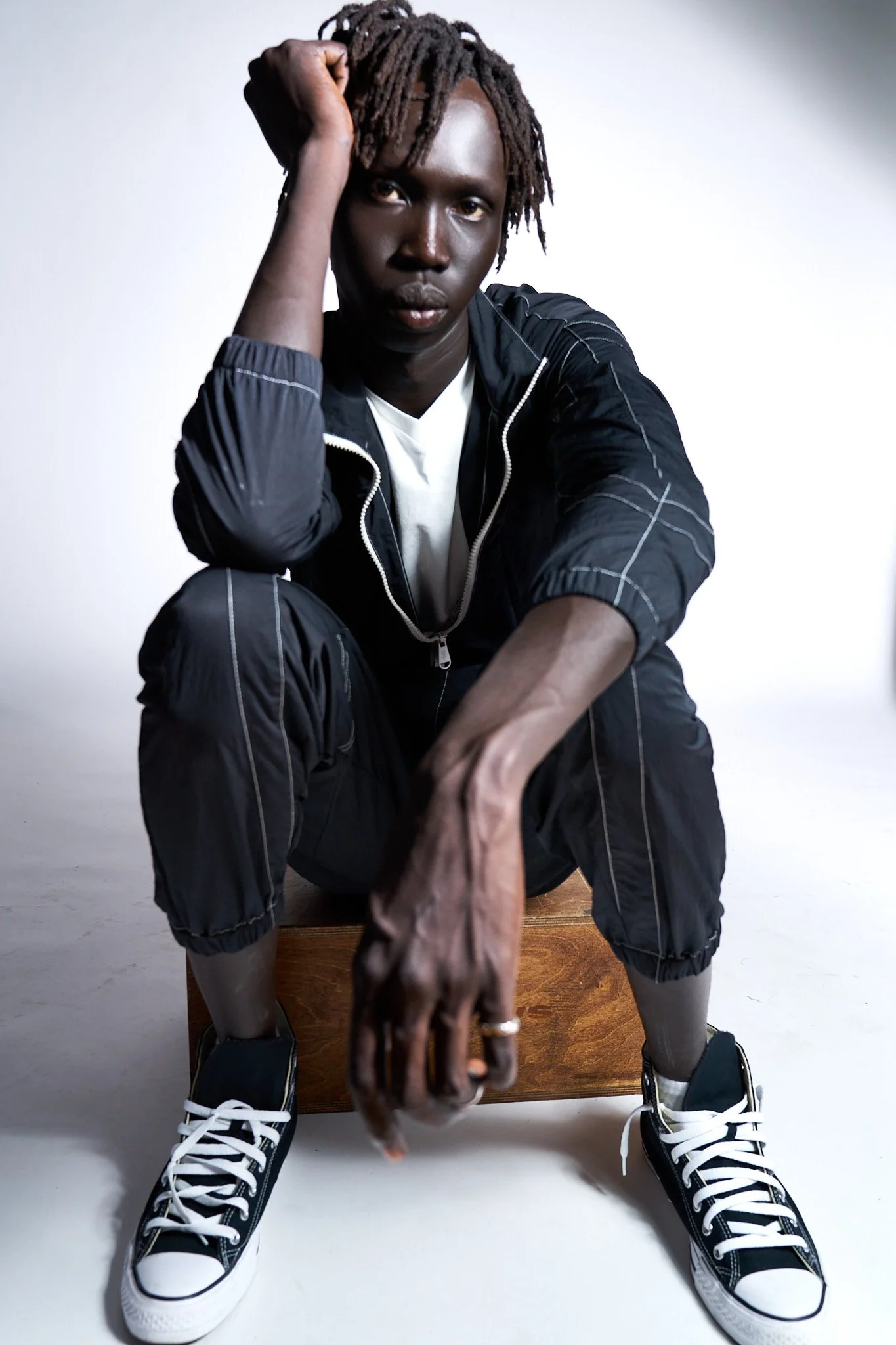 A person with dreadlocks, wearing a black jacket, white shirt, black shorts, and black sneakers with white laces, sitting on a wooden box in front of a white background, reaching forward with a hand.