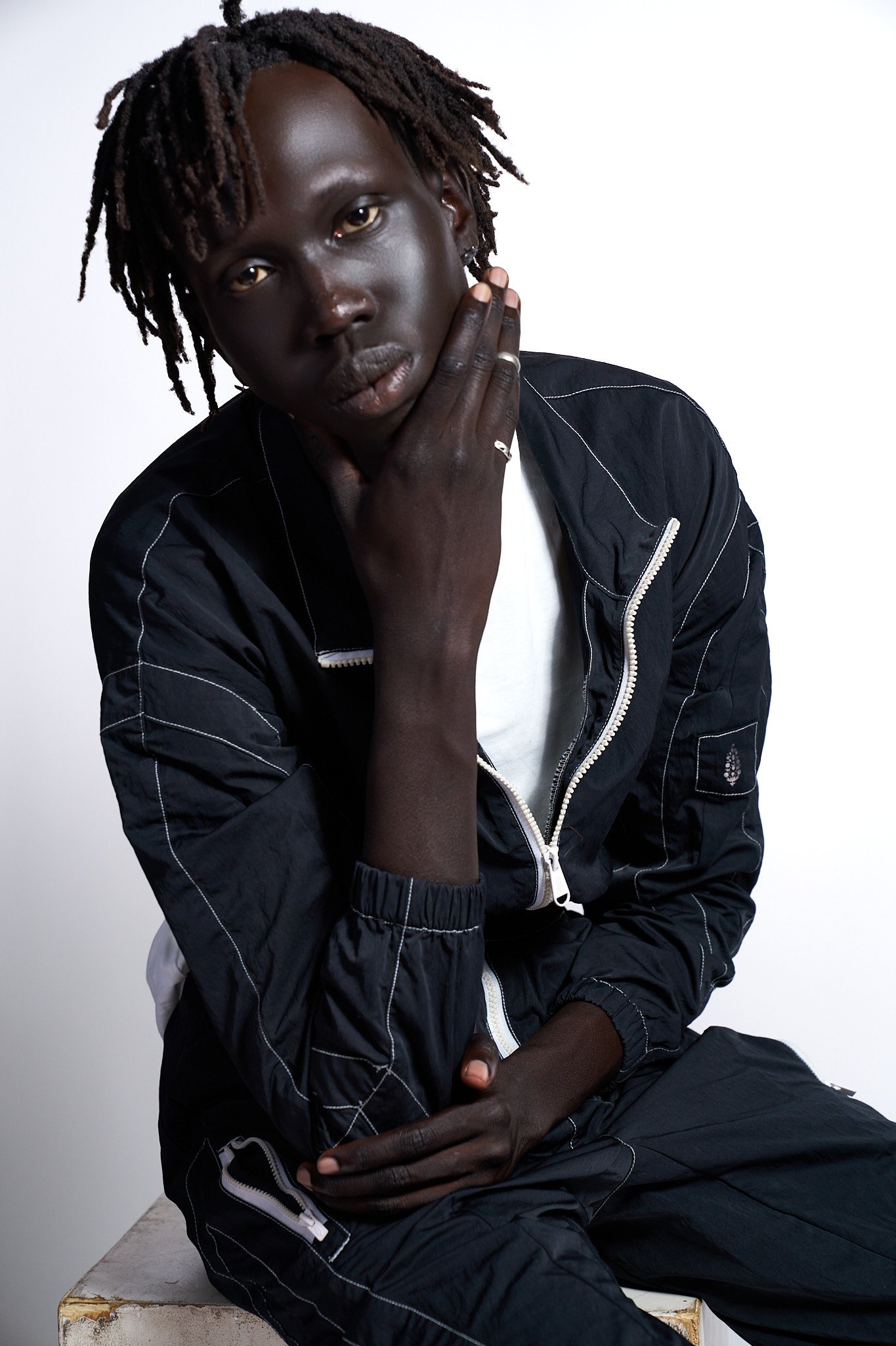 A young man with dark skin and dreadlocks poses against a plain white background, wearing a black bomber jacket and white shirt, with his hand resting on his face.