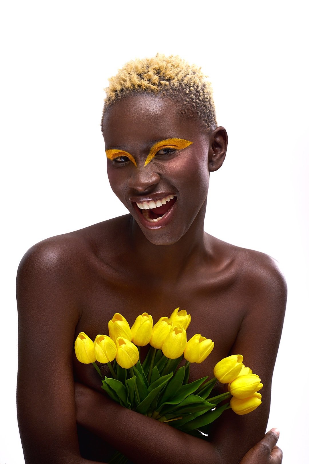 A smiling woman with short, curly blonde hair, bold yellow eye makeup, holding a bouquet of yellow tulips, standing against a plain white background.