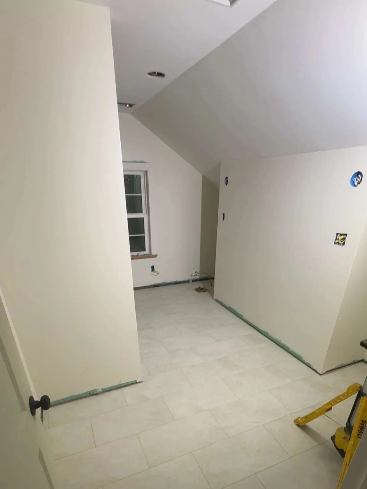 An interior room under construction with white walls and light-colored tiled flooring. There is a window on the back wall, an open doorway on the left, and some construction tools and materials. The ceiling has recessed lighting fixtures being installed.