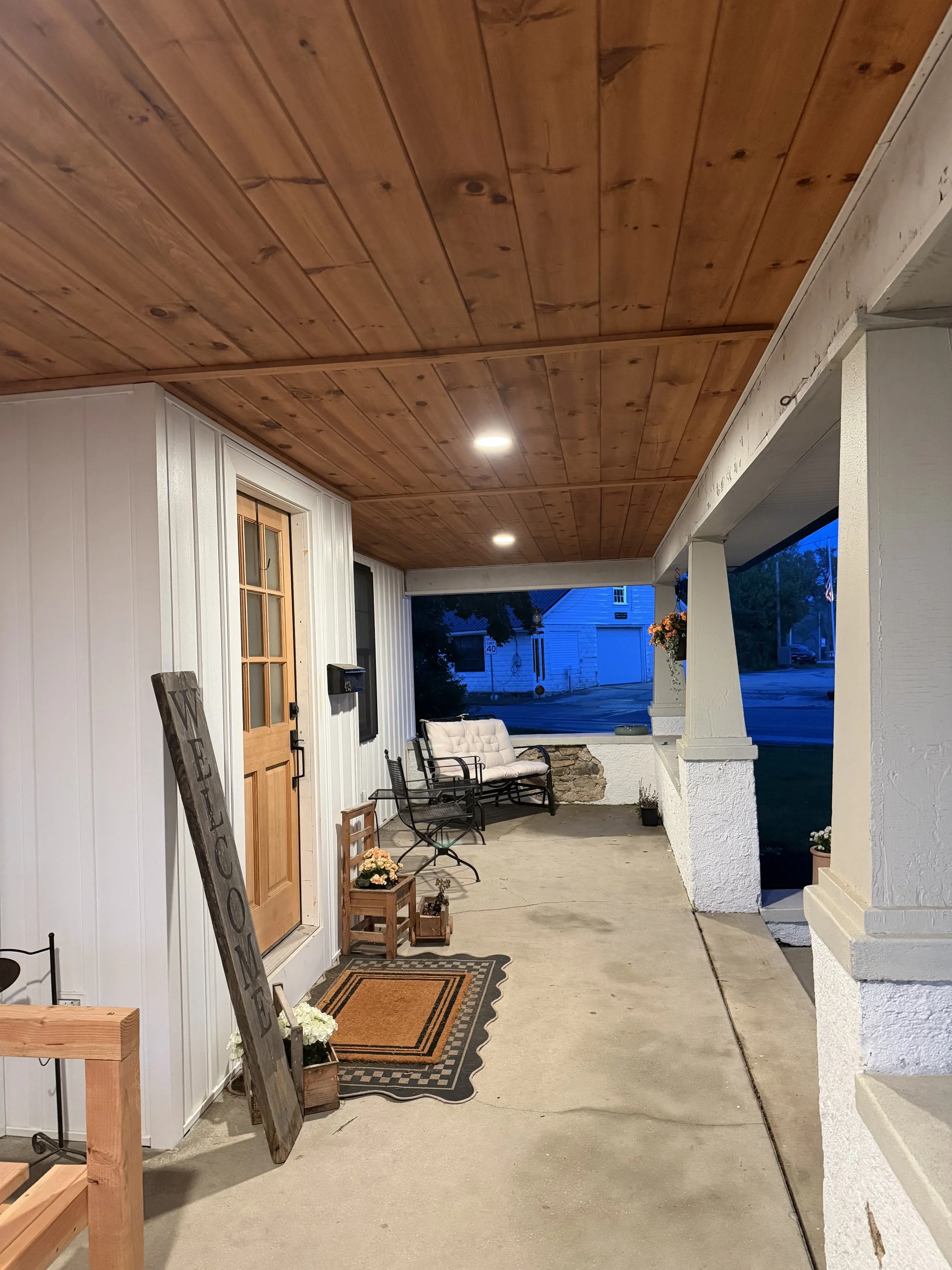 Covered porch area with a wooden ceiling, white walls, porch furniture including a cushioned bench and chairs, potted plants, a door with a window, and decorative items near the entrance at dusk.