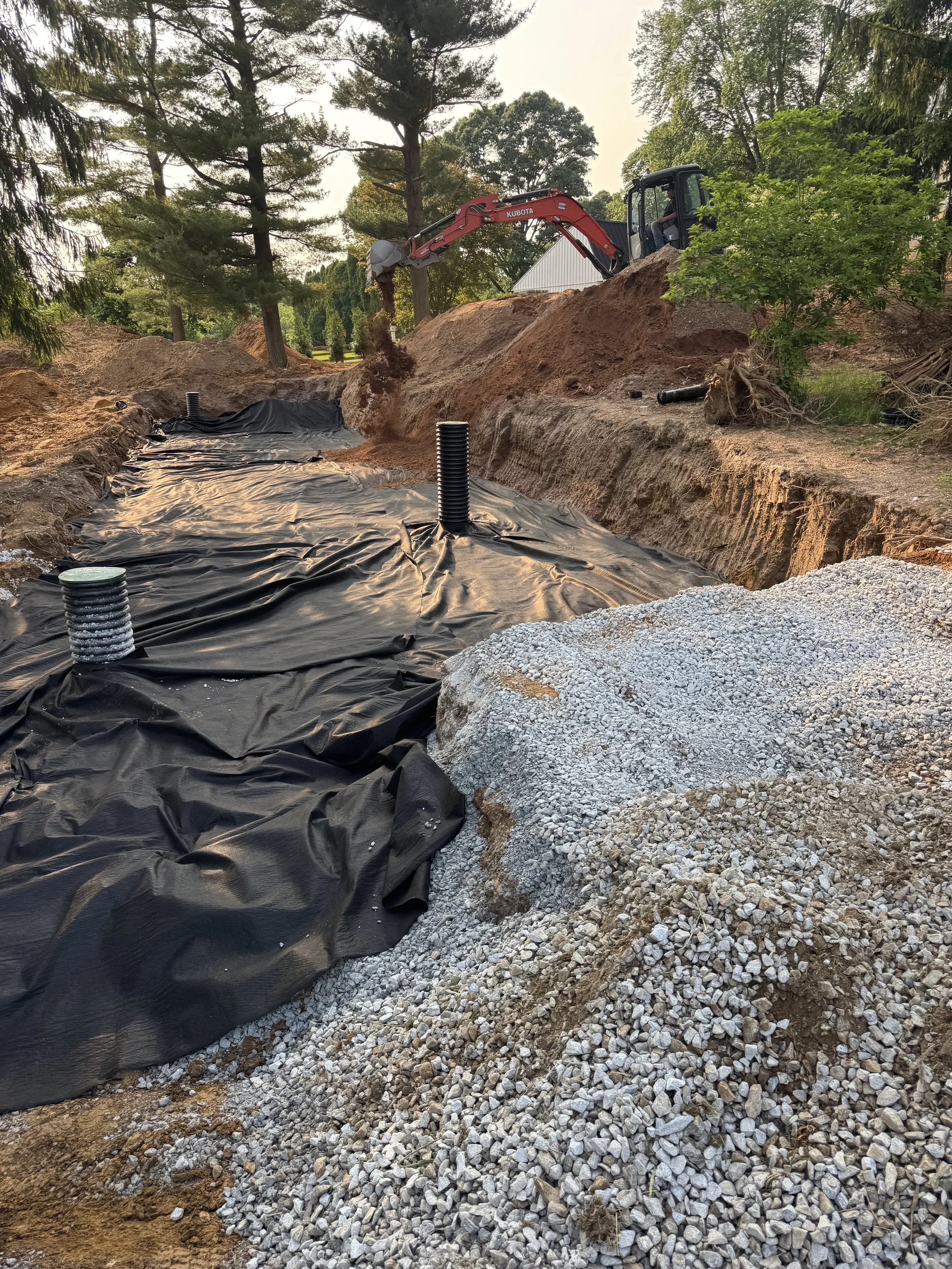 A construction site with a small excavator digging a trench. The trench is lined with black plastic sheeting and vent pipes sticking up through it. There are piles of gravel and dirt surrounding the trench, and trees and a building are visible in the background.
