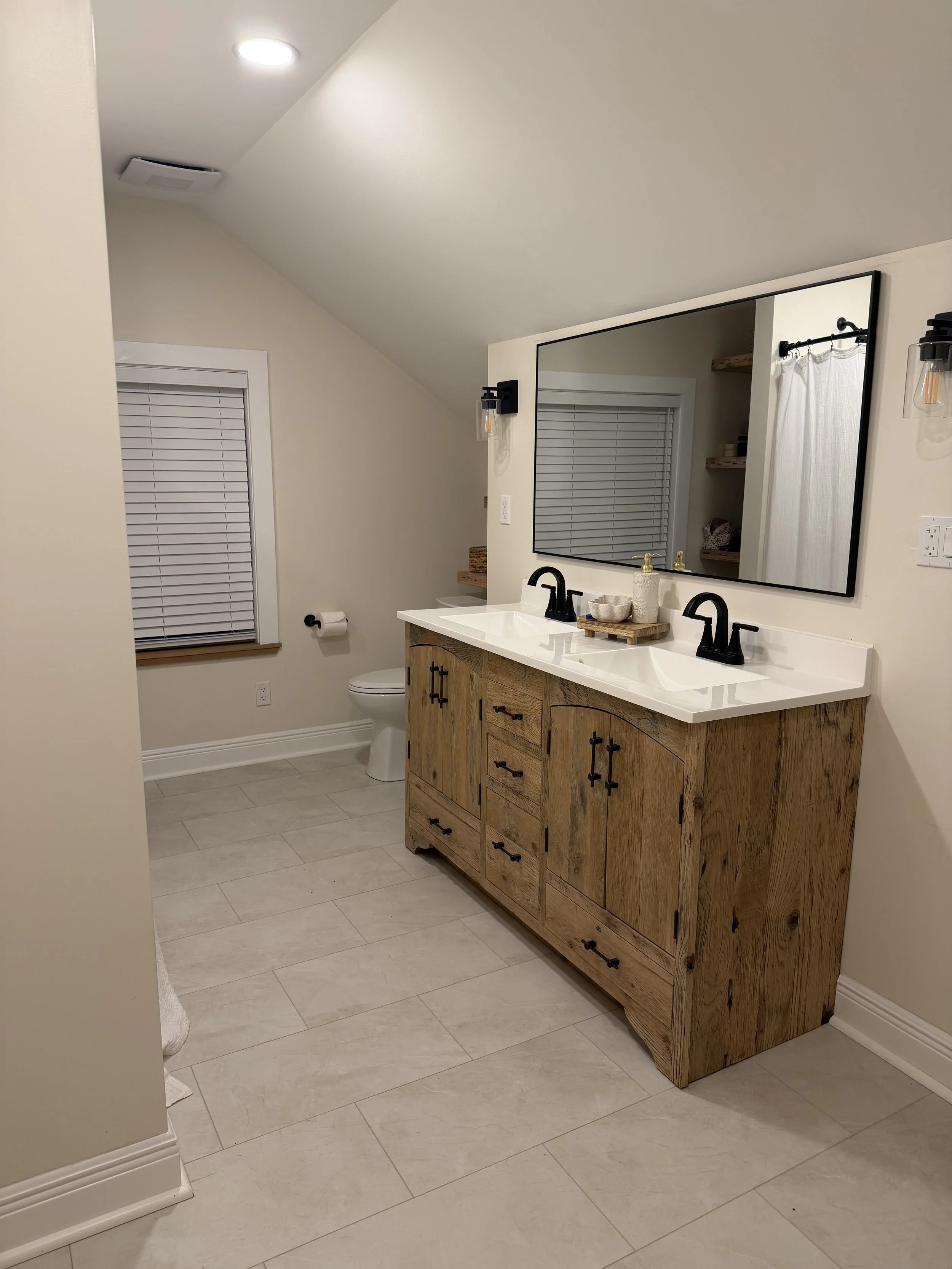 A bathroom with a wooden vanity, a large mirror, black faucets, and a white countertop. There is a window with white blinds, a toilet, and small floating shelves with baskets. The floor is tiled in light color.