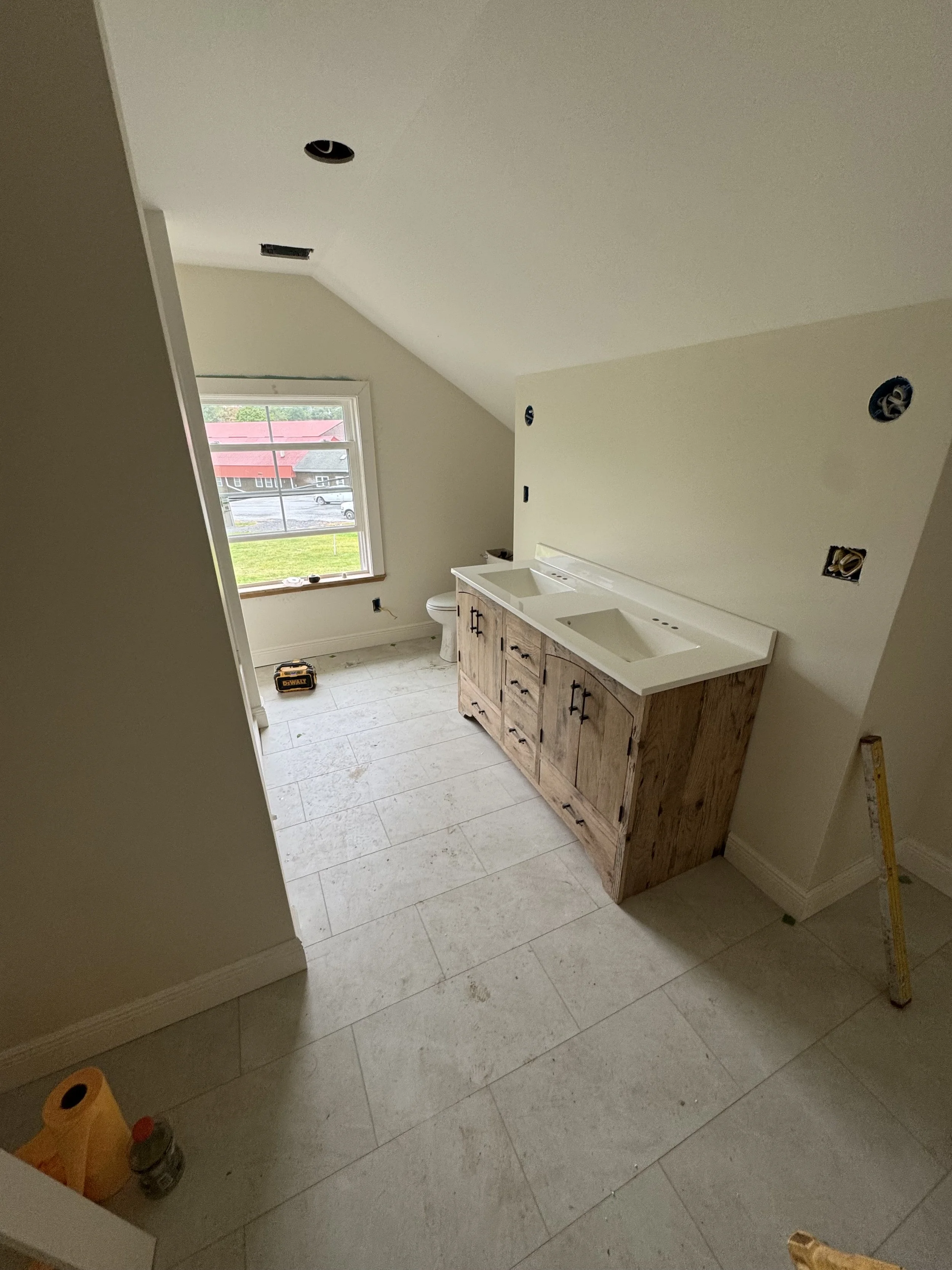 A bathroom under construction with a double sink vanity, wall outlets and electrical boxes, a window showing a red roof outside, and tools on the floor.