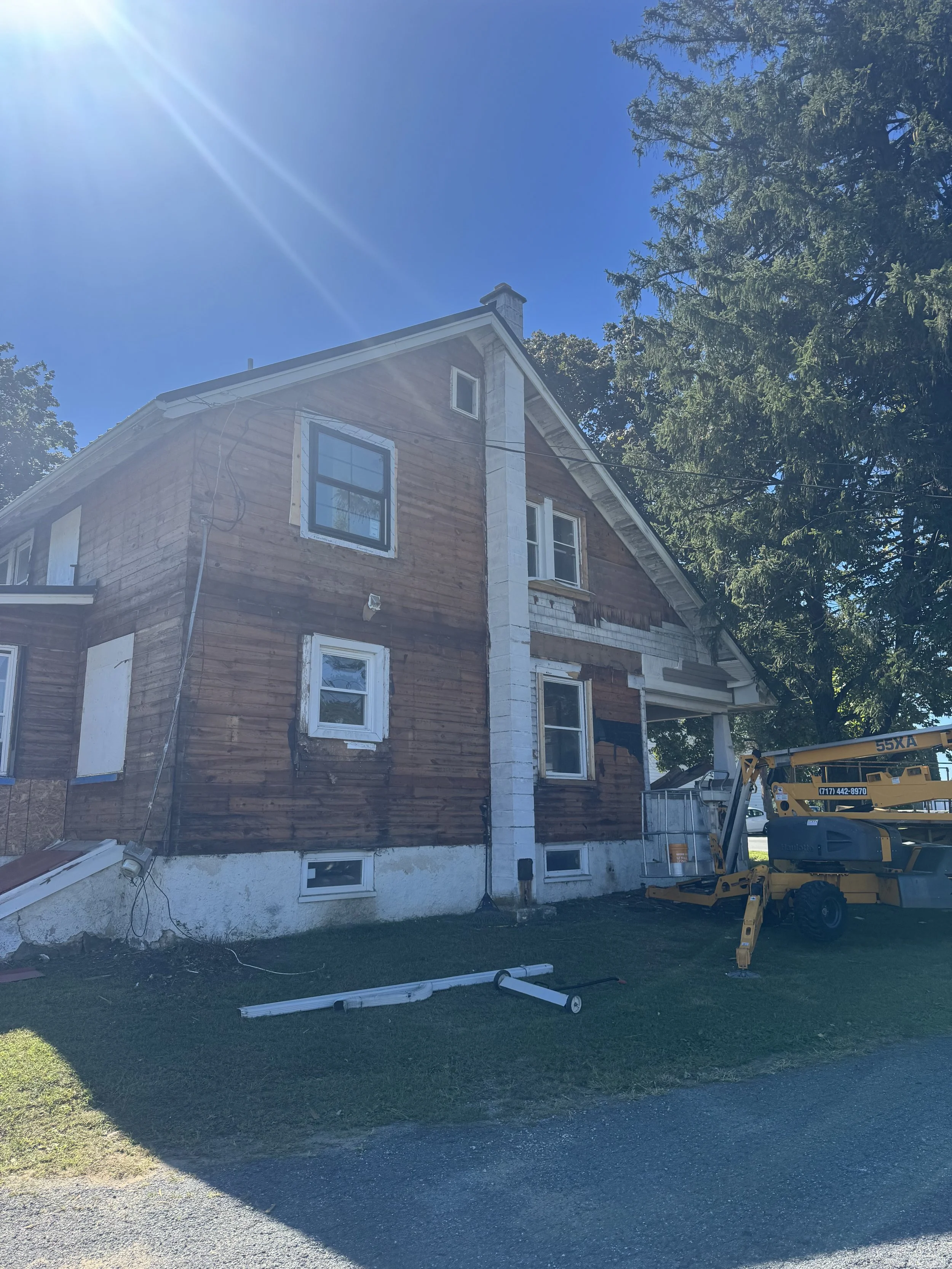 House under renovation with siding partially removed, construction equipment nearby, and debris on the ground, under a bright sunny sky.