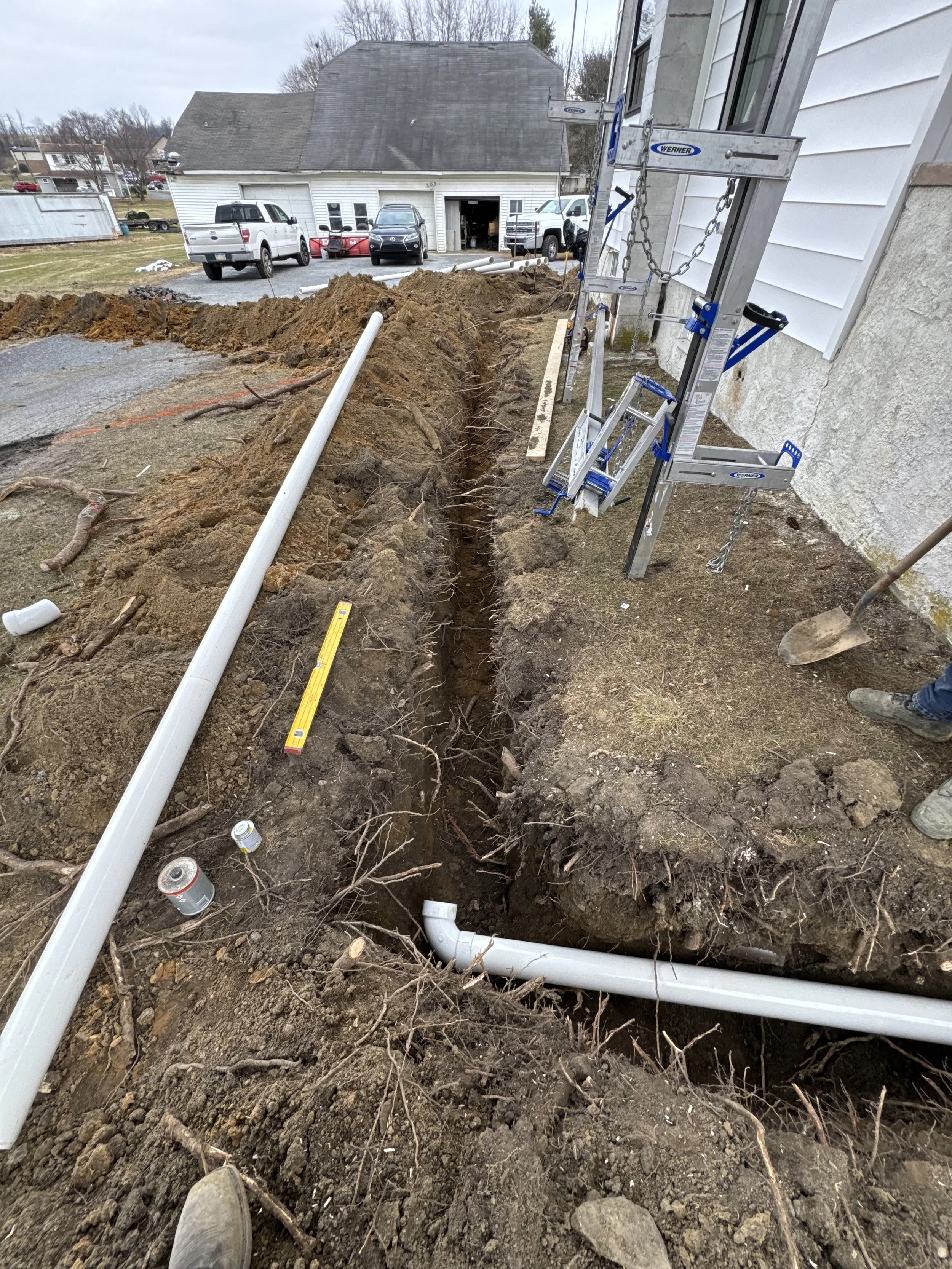 Underground plumbing installation in progress next to a house, with pipes, dirt, and construction tools visible.