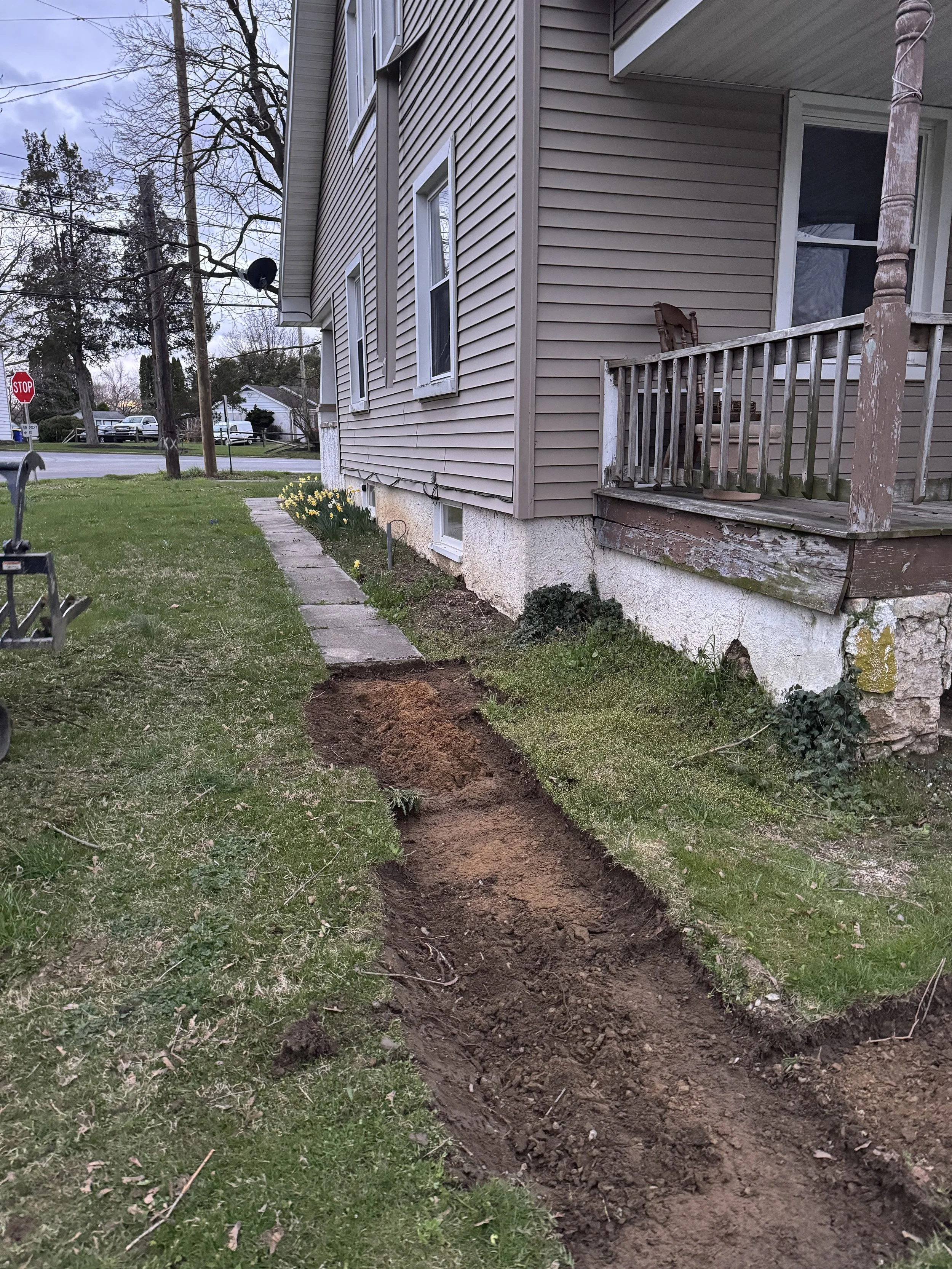 A narrow trench dug into the grass yard along the side of a house, with a concrete walkway next to it. The house has beige vinyl siding, white trim, and a small porch with a wooden railing and an unfinished wooden post. In the background, there are trees, a street with a stop sign, and a few parked cars.