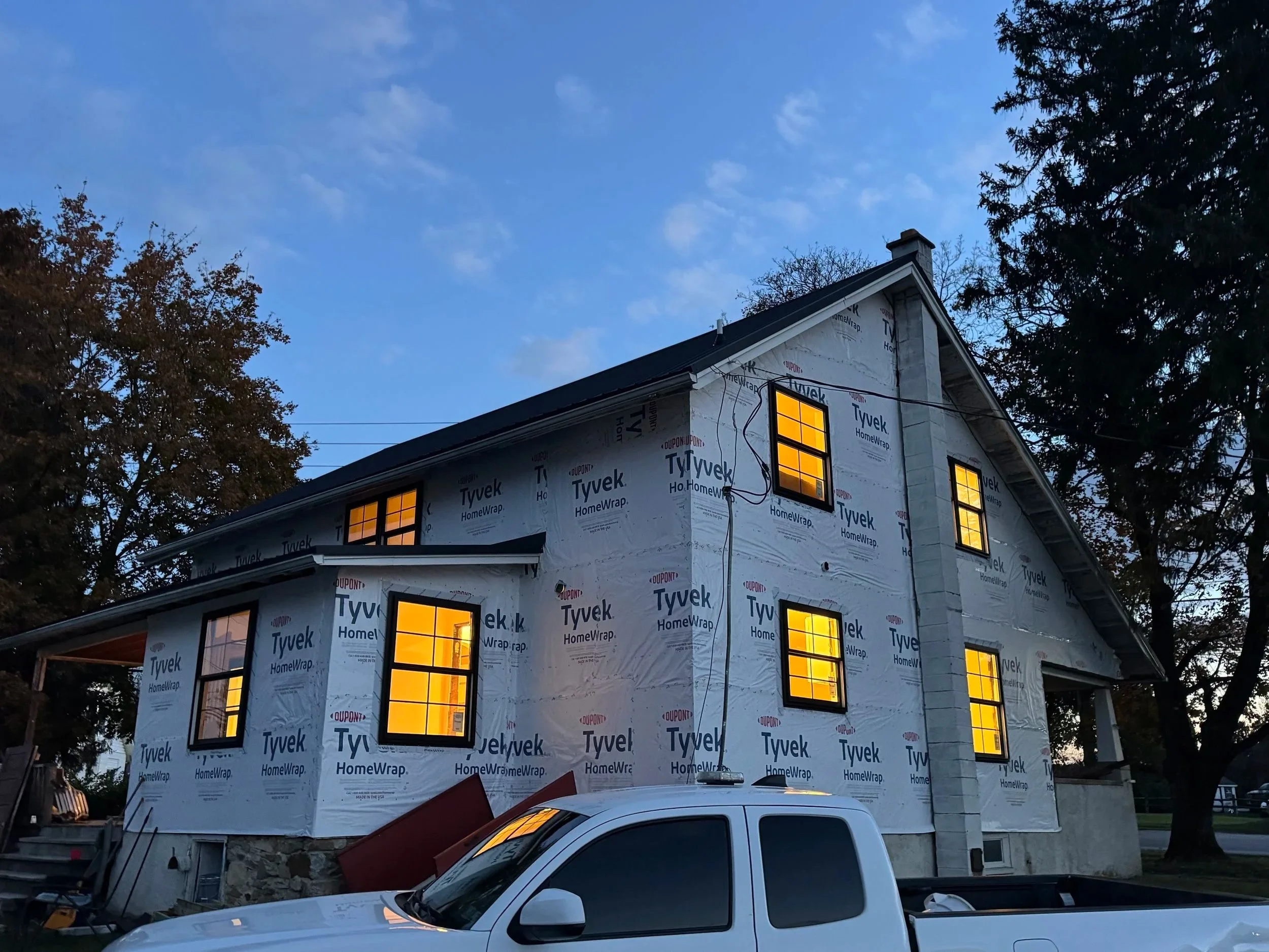 House under construction wrapped in Tyvek HomeWrap with illuminated windows and a dark evening sky.