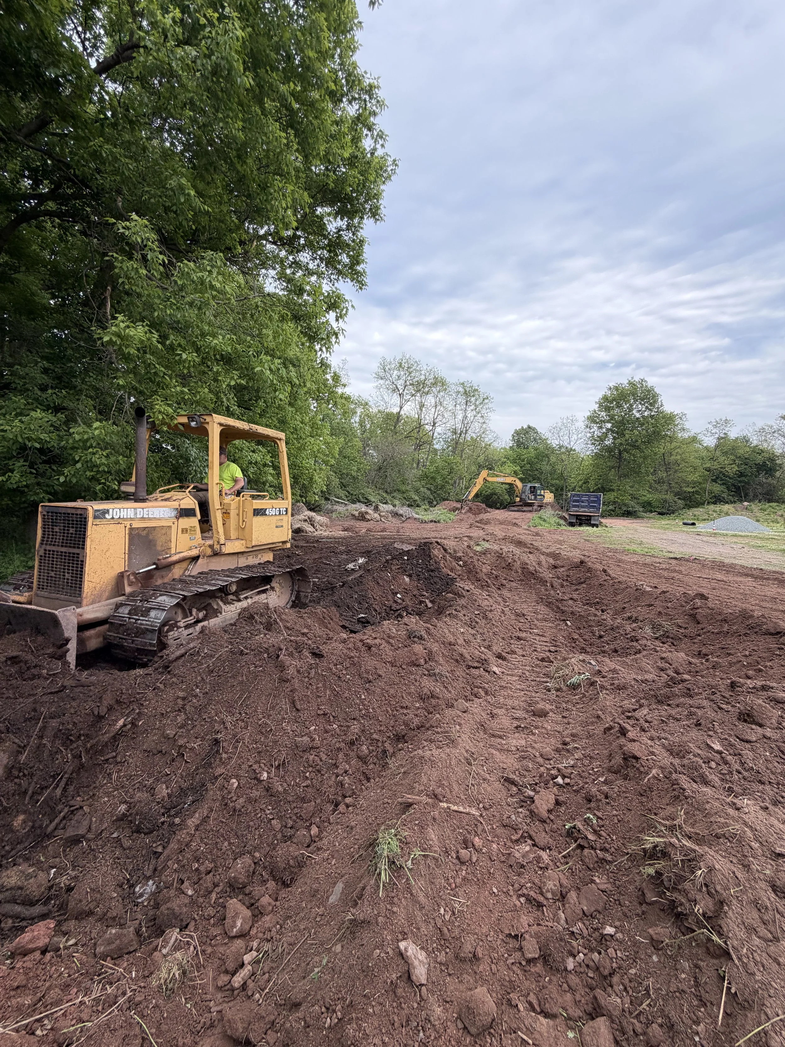 Construction site with a John Deere bulldozer on dirt, trees and cloudy sky in the background.