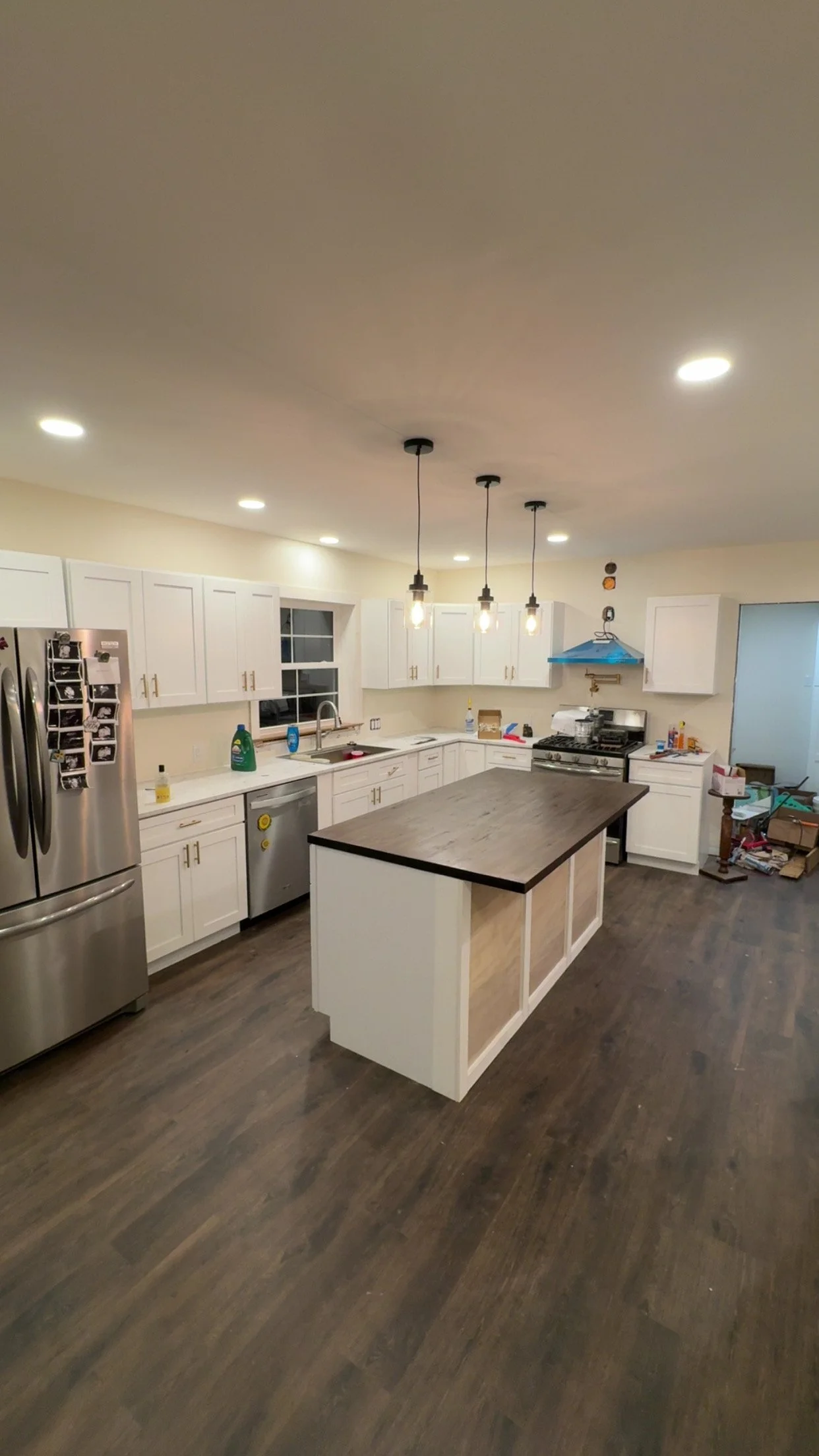 Kitchen with white cabinets, a central island with a dark countertop, stainless steel appliances including a refrigerator and a stove, and a set of three pendant lights hanging from the ceiling. The room appears to be under renovation or cluttered with some boxes and items on the countertops and floor.