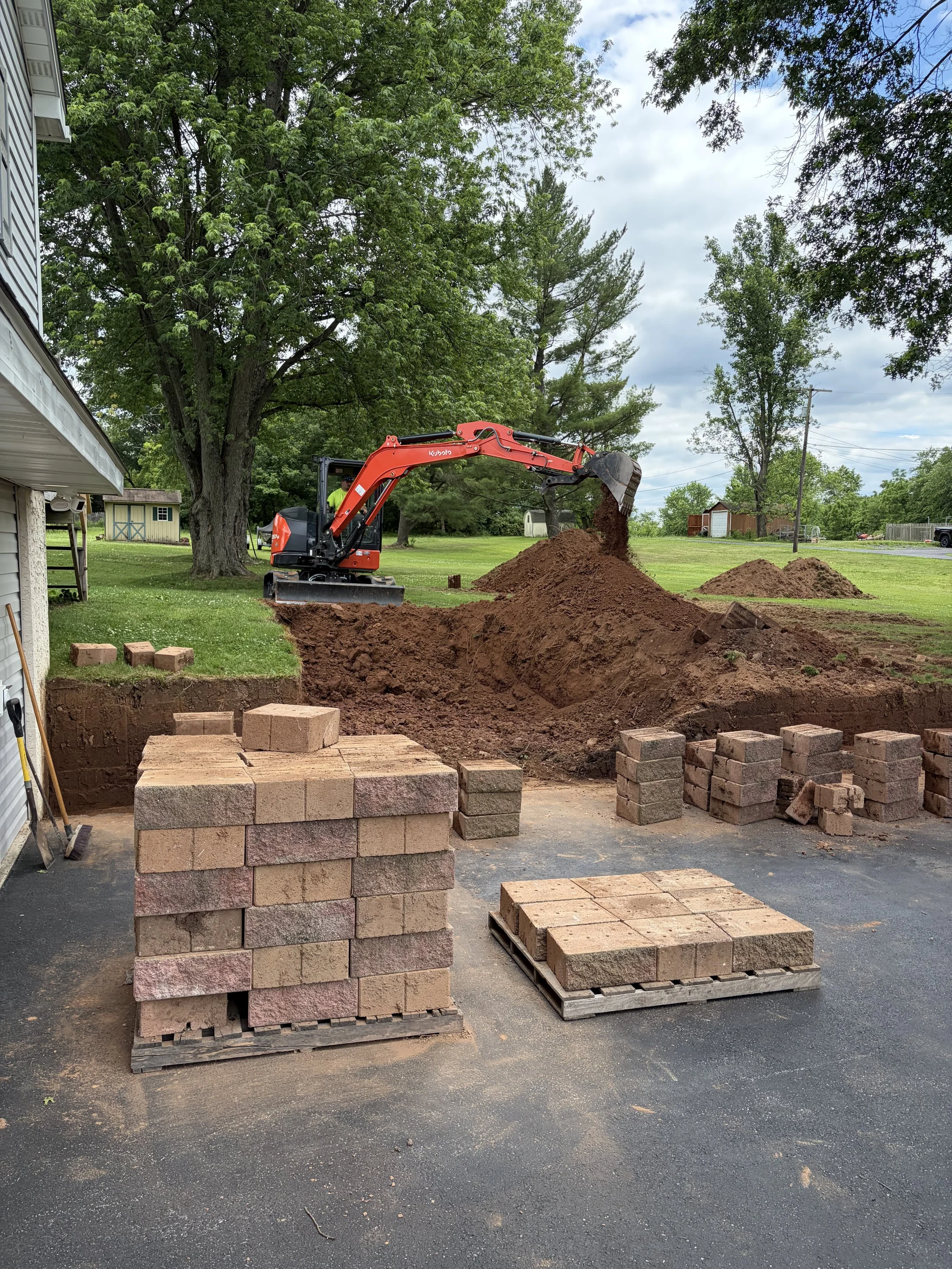 A backyard with a small excavator pouring dirt into a trench. Several stacks of bricks are on pallets nearby. Large trees and a cloudy sky are in the background.