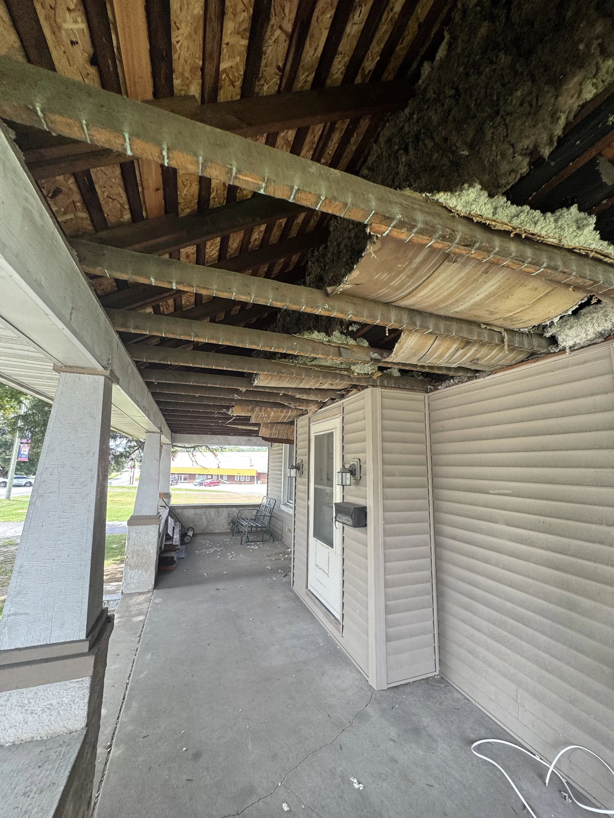 The underside of a porch roof with exposed wooden beams and insulation materials, with some debris on the concrete floor, and part of the house's siding visible.