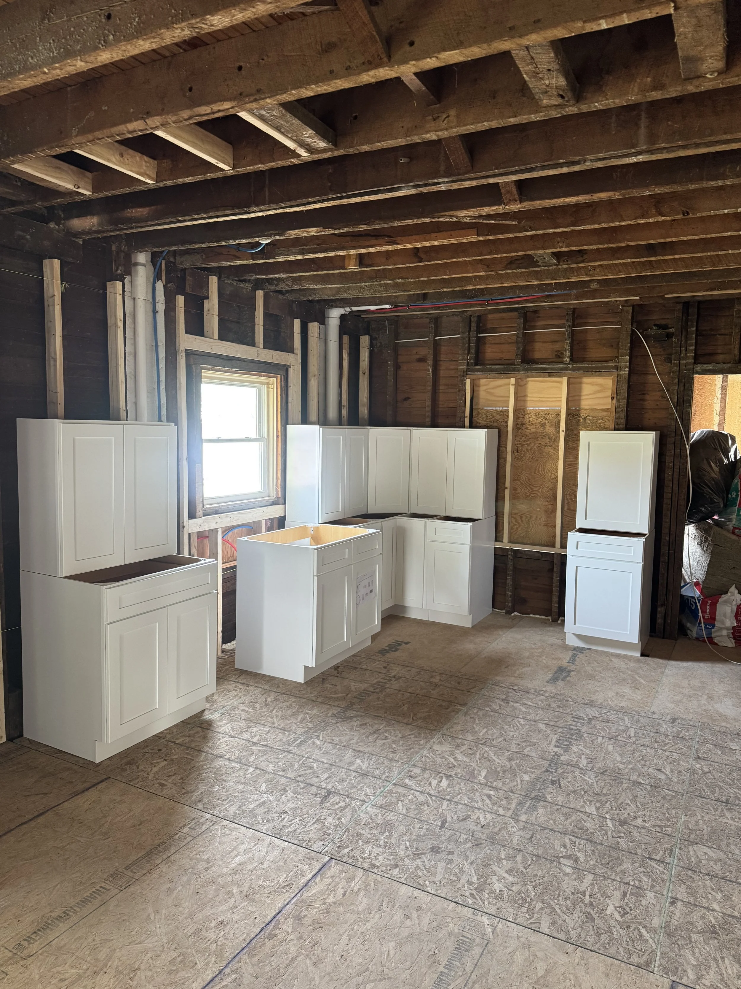 Interior view of a room under construction with exposed wooden ceiling beams, partially finished walls, and white kitchen cabinets installed.