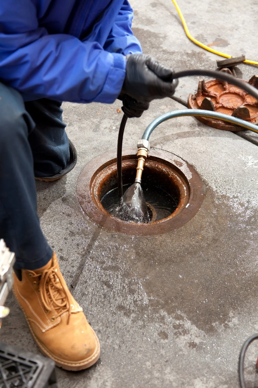 A person in blue clothing is working on a sewer pipe, using a tool connected to a hose that is inserted into an open clean-out. The person is wearing black gloves and tan work boots, and there is a rusty pipe cover nearby.