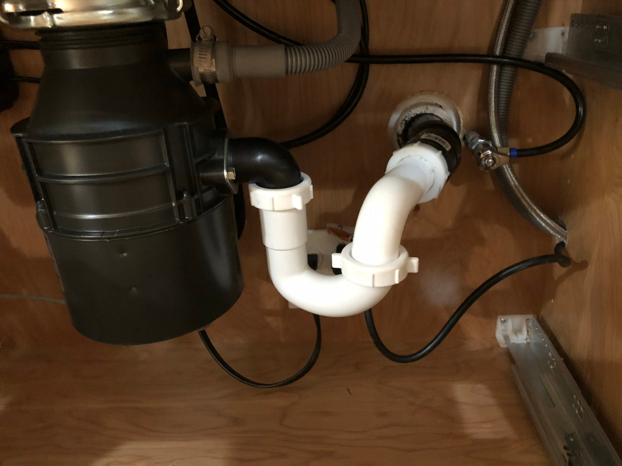 Underneath a kitchen sink with pipes, hoses, and a black disposal unit, installed on a wooden cabinet floor.