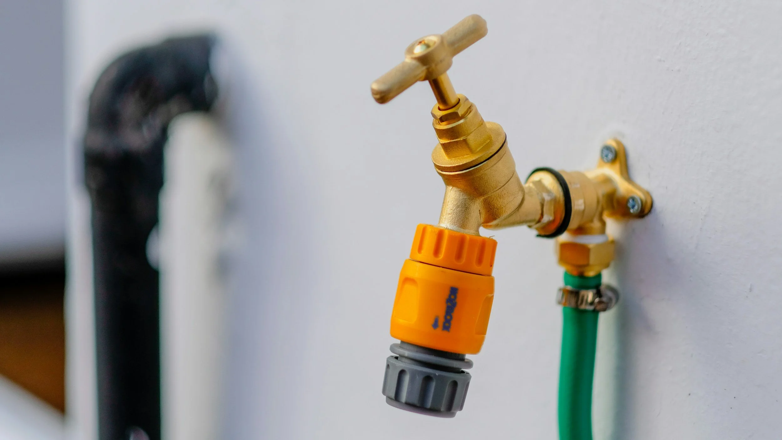 Close-up of a brass water faucet attached to a white wall, with a yellow gardening hose connector and green and black hoses connected.