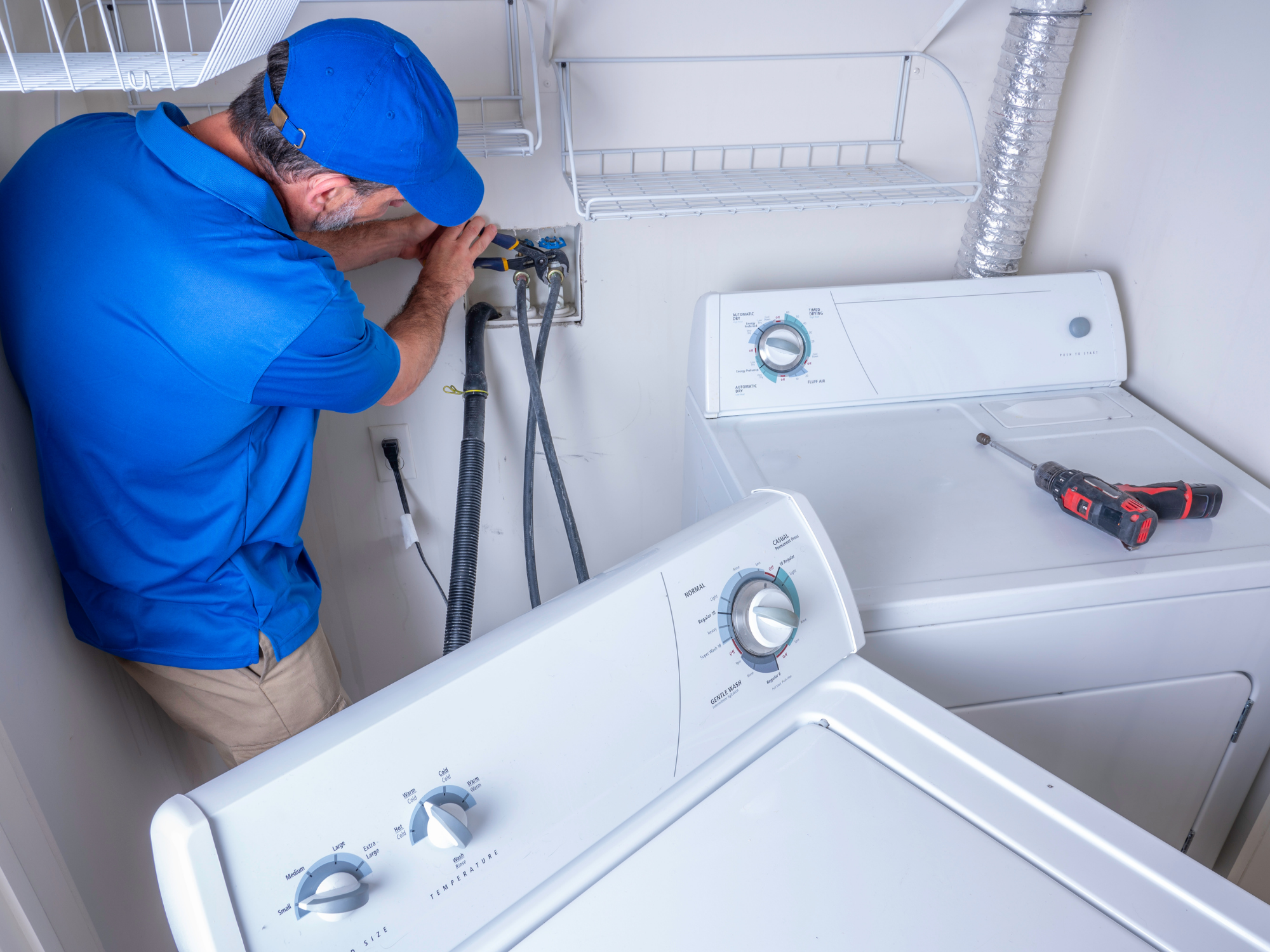 A person repairing a washing machine in a laundry room. The individual is wearing a blue cap and shirt, and using tools to fix the washing machine.