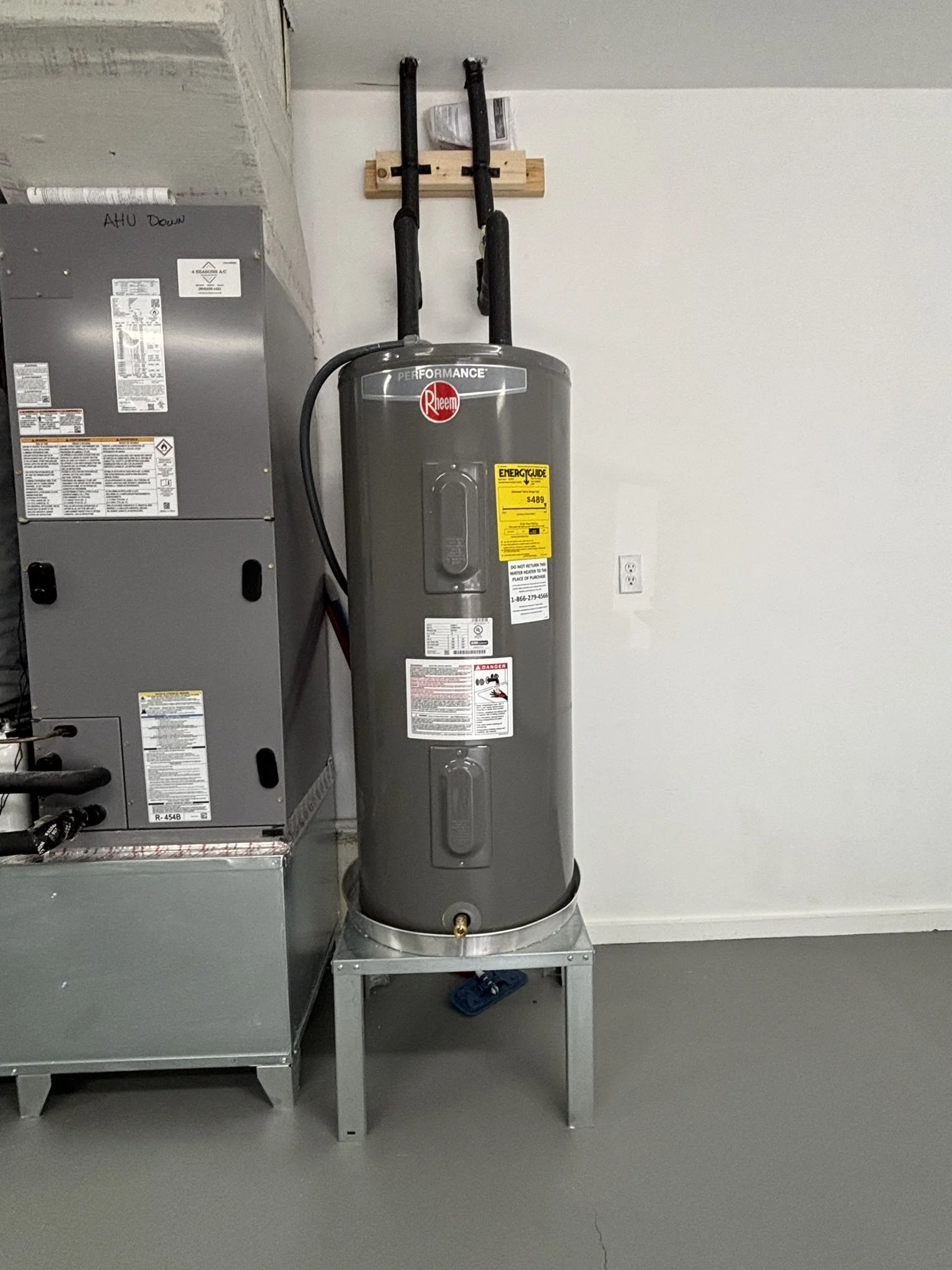 A water heater installed on a small metal stand in a utility room, with pipes on top and a yellow energy guide label on the front.