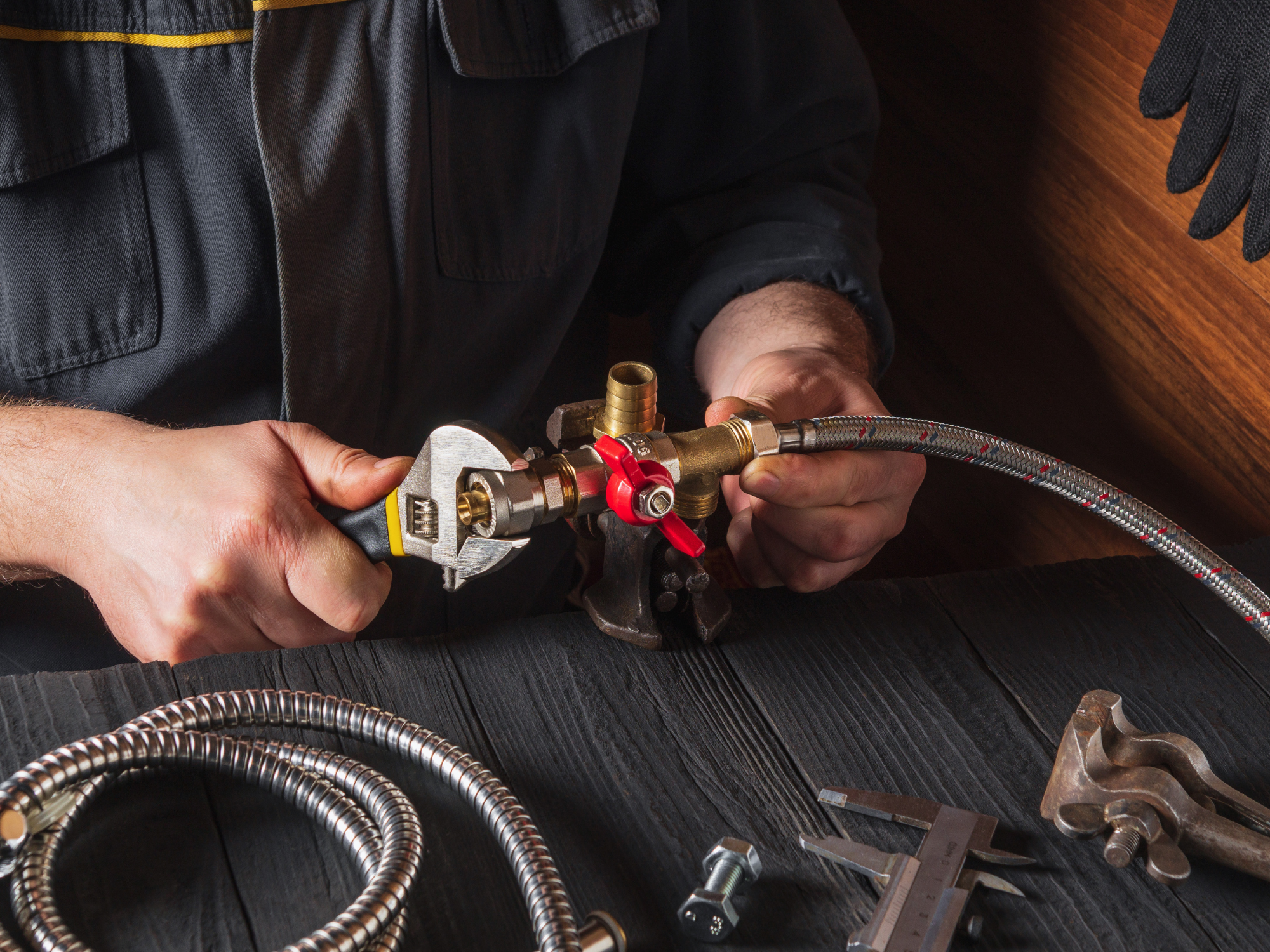 A person wearing black clothing and gloves working with plumbing tools, holding a pipe fitting and adjusting a brass valve connected to flexible hoses on a wooden surface.