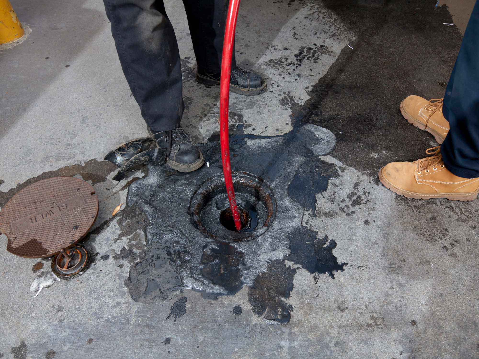 Construction workers' legs and feet around an open manhole on a city street. One worker is near the manhole with a red tool, others wearing work boots standing nearby. Wet asphalt and a traffic bollard are visible.