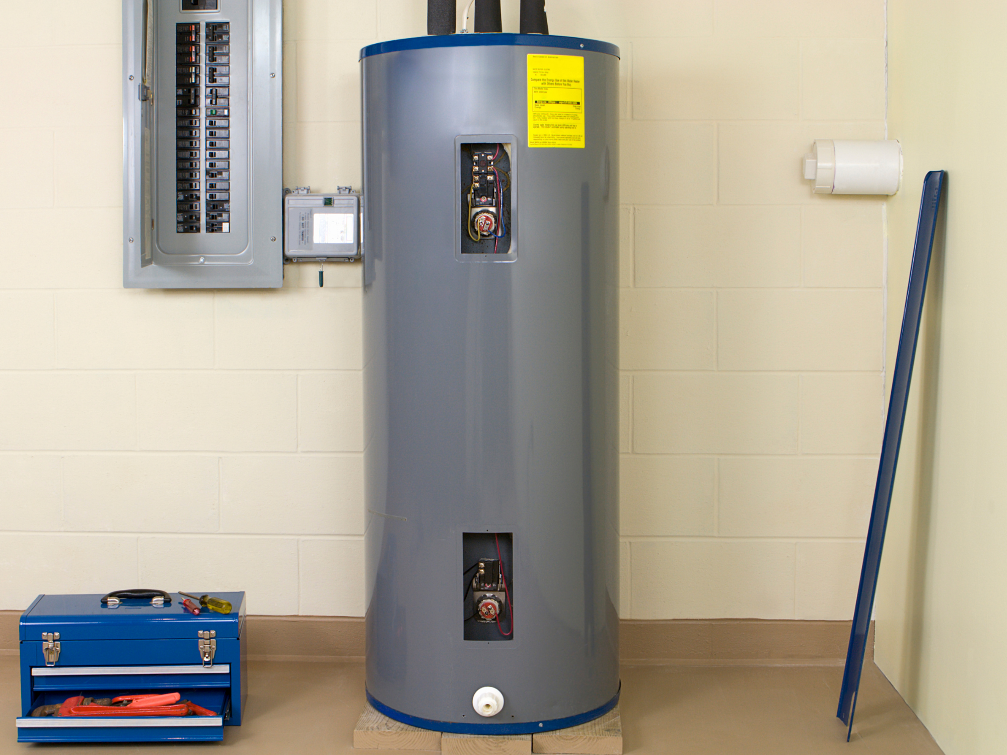 A water heater tank with its front panel removed, exposing wiring and components inside. A blue toolbox and a set of screwdrivers and tools sit on the floor in front of the tank. An electrical panel is on the wall to the left and a white pipe on the wall to the right.